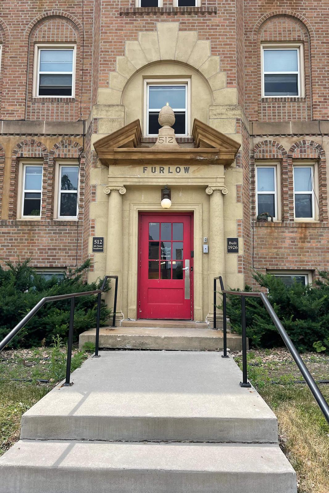 Facade of a brick apartment building with a prominent entrance. The door is painted bright red, flanked by decorative stone features. Above the door, the name "FURLOW" is carved into the stone. A concrete staircase leads up to the entrance, surrounded by green shrubs and grass.