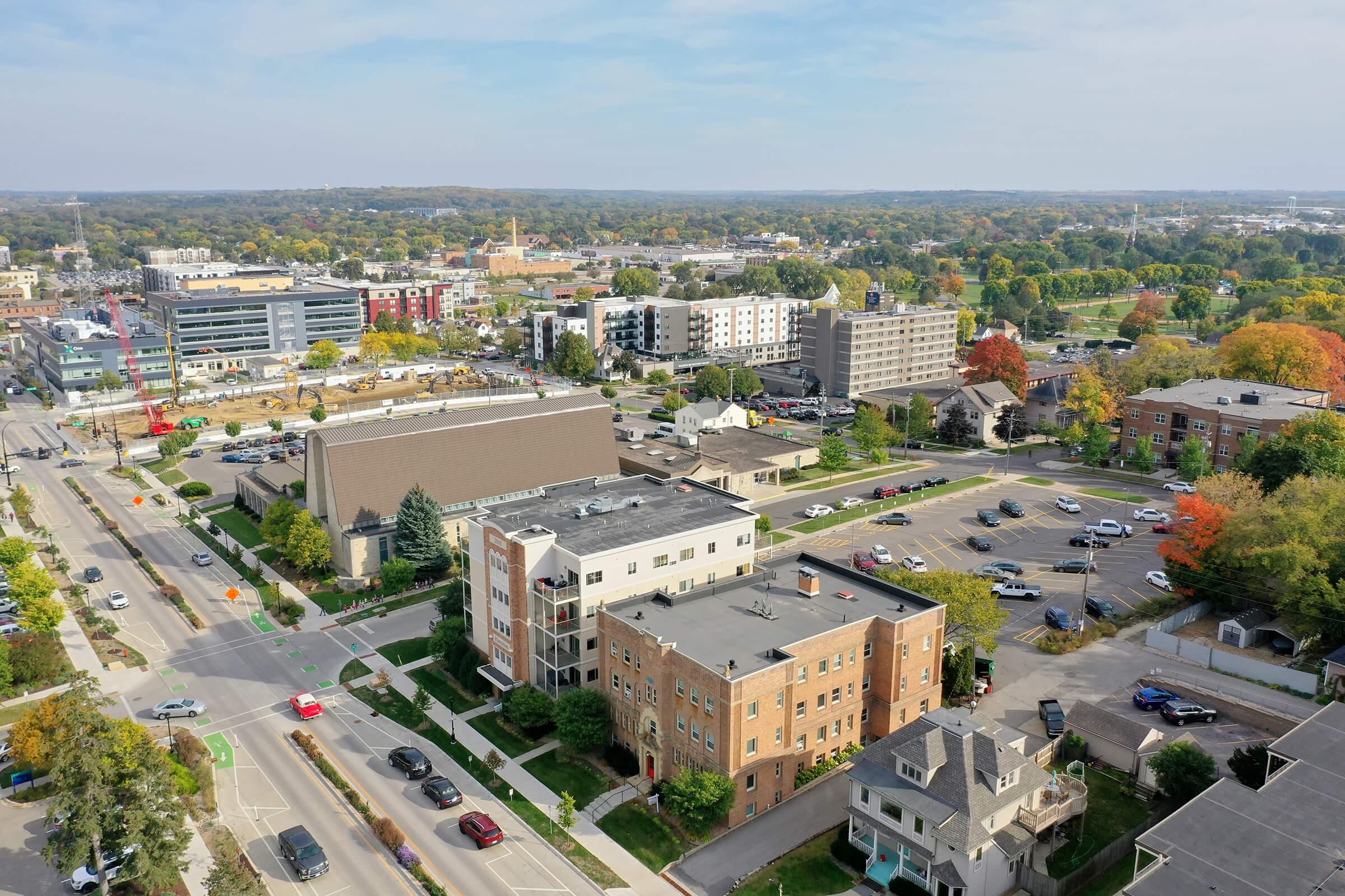 Aerial view of a suburban area featuring a mix of residential and commercial buildings. Trees with autumn foliage line the streets, and several cars are parked along the roads. In the background, various structures including shops and apartments can be seen, set against a clear blue sky.
