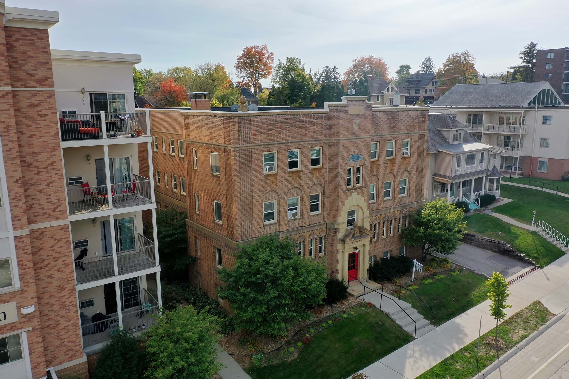 Aerial view of a brick building with large windows and a red door, surrounded by greenery. Nearby, there are modern apartment buildings with balconies, set against a backdrop of trees showing autumn colors. A pathway leads to the entrance of the brick building.