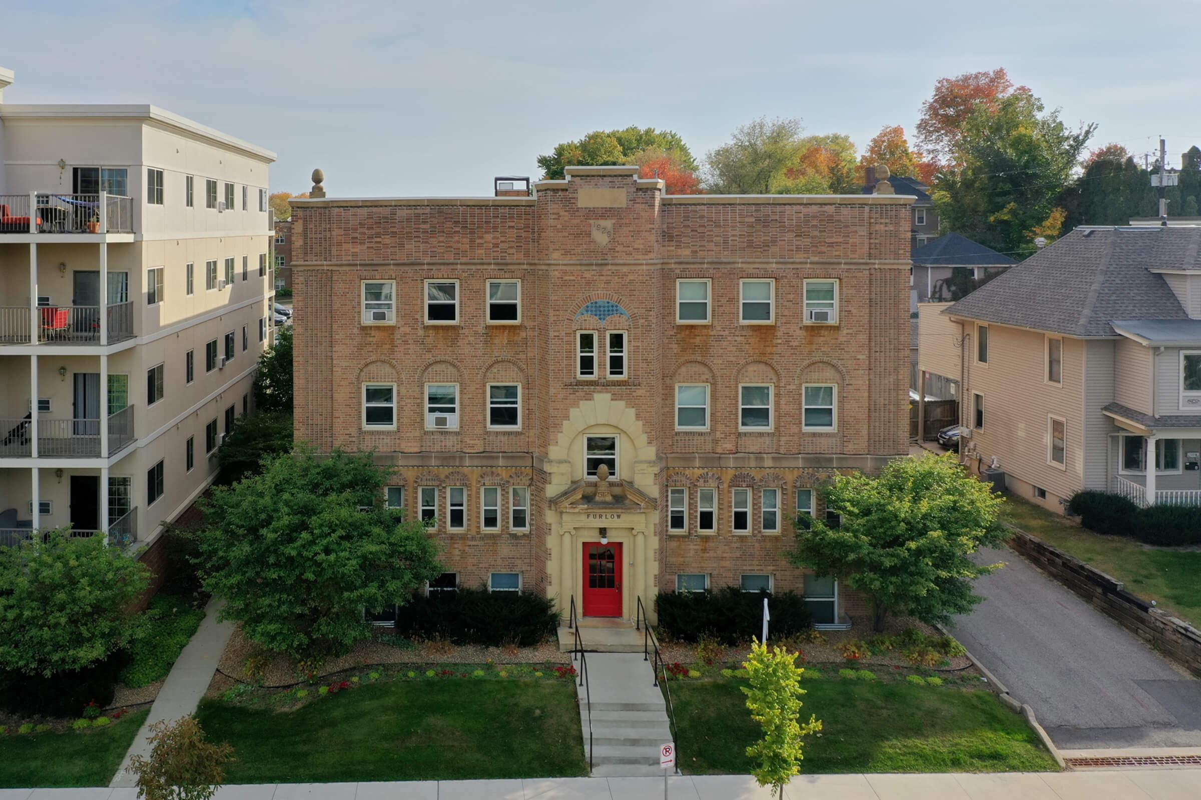 A historic brick building featuring a stone entrance and decorative architectural elements, surrounded by trees and small shrubs. Nearby, there are modern apartment buildings visible on the left. The scene is set under a clear sky with hints of autumn foliage in the background.