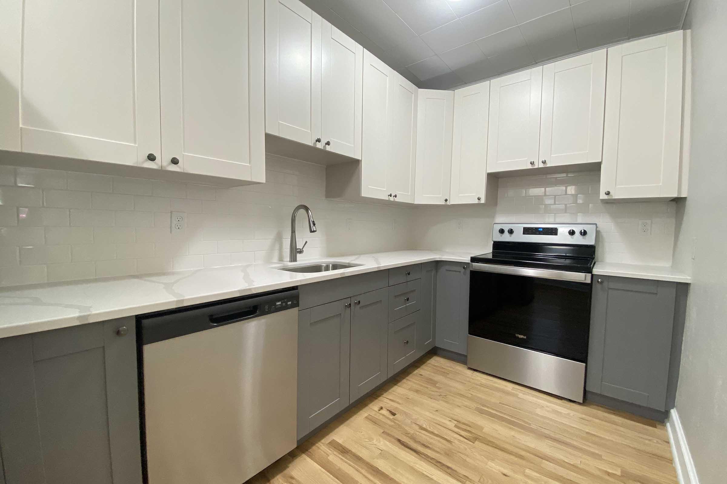 Modern kitchen featuring a corner layout with light gray and white cabinets, stainless steel appliances including a dishwasher and stove, a stainless steel sink, and a white tiled backsplash. The floor is made of natural wood, adding warmth to the contemporary design.