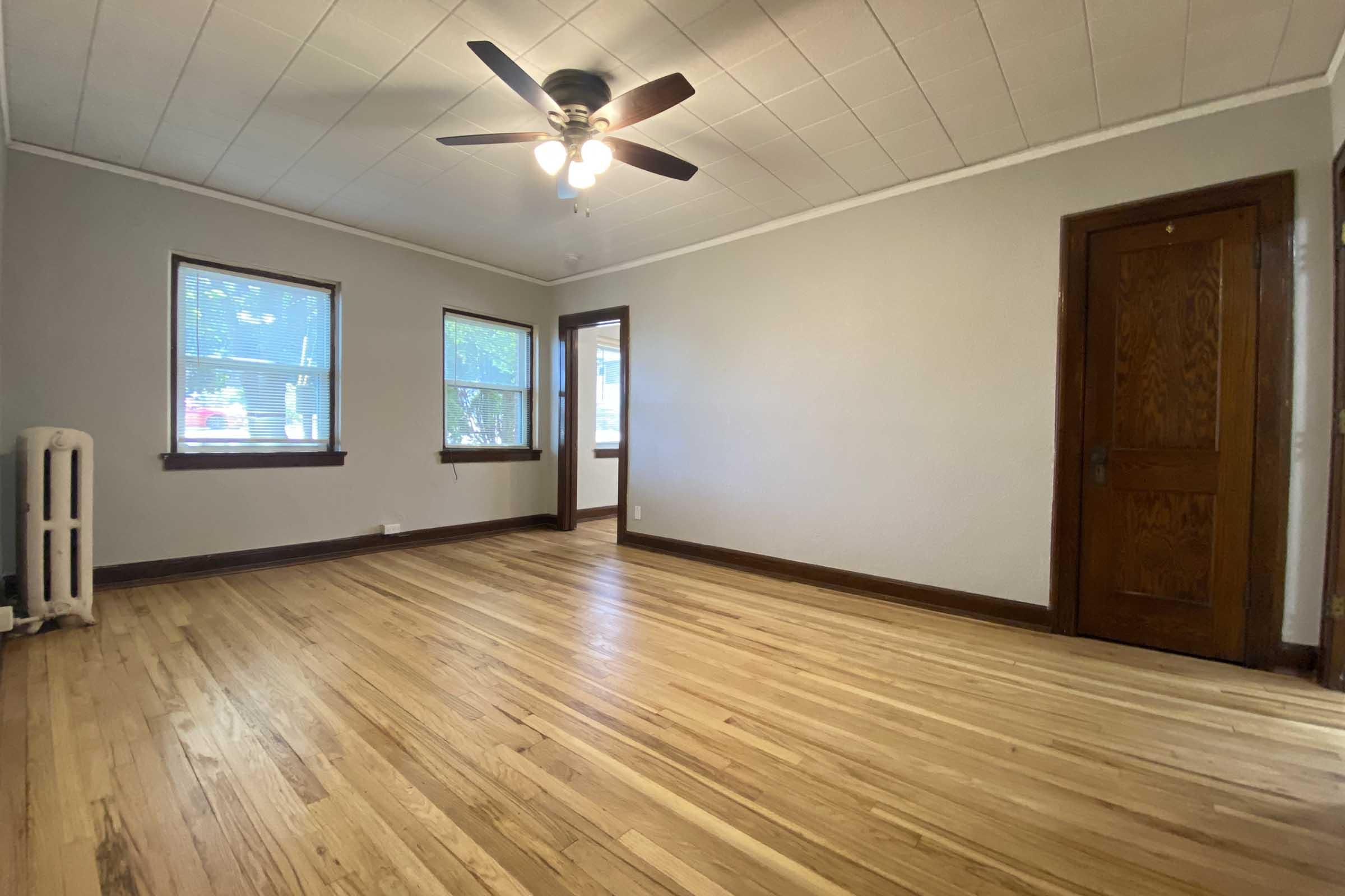 A spacious, empty room with hardwood flooring, featuring a ceiling fan with light fixtures, two windows allowing natural light, and a wooden door. A vintage-style radiator is visible in the corner, adding character to the otherwise bare space. The walls are painted a light color, enhancing the room's brightness.