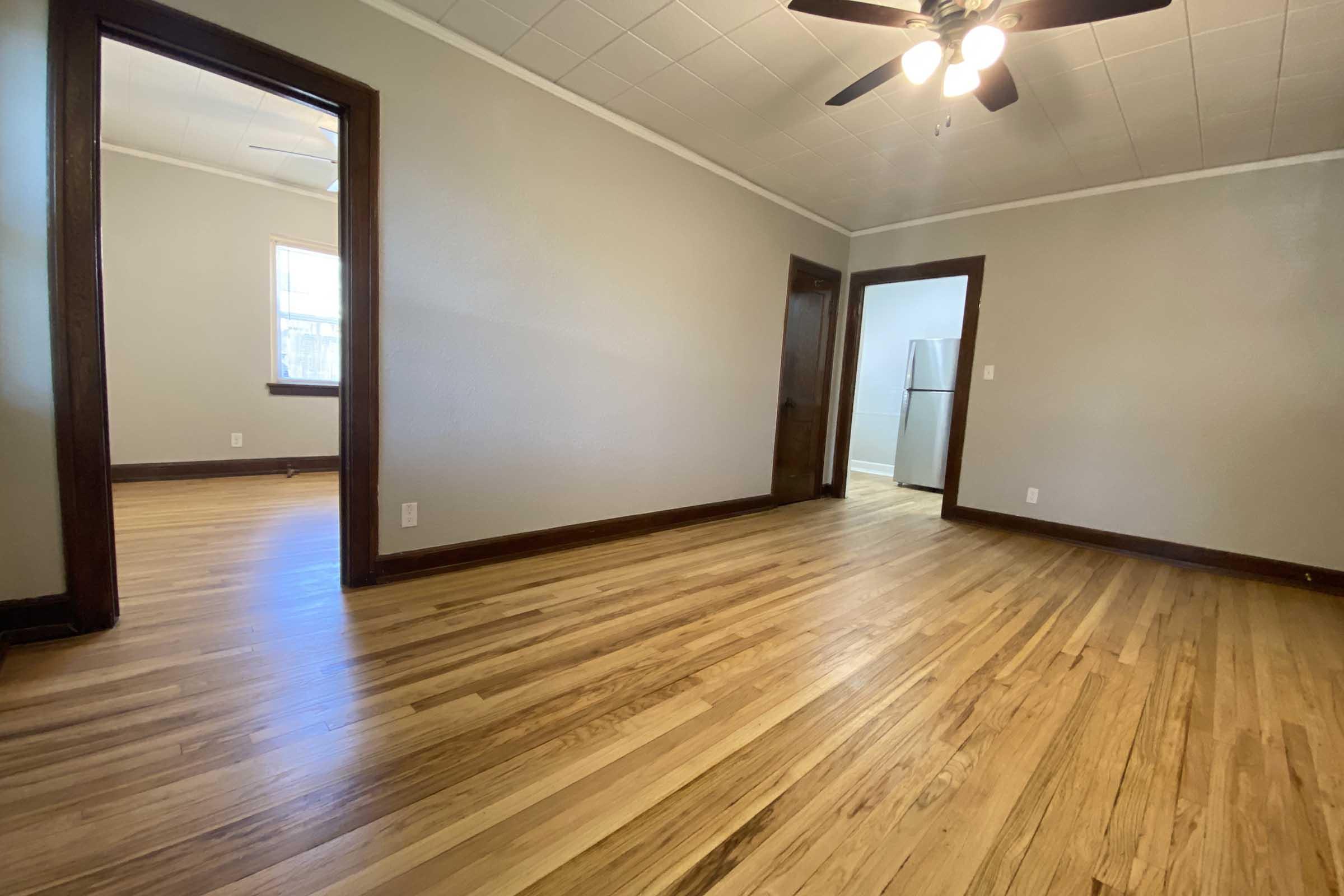 A spacious, empty room featuring hardwood floors, light gray walls, and a ceiling fan. There's a doorway leading to another room and a glimpse of a kitchen area in the background. Natural light comes through a window, creating a bright and inviting atmosphere.