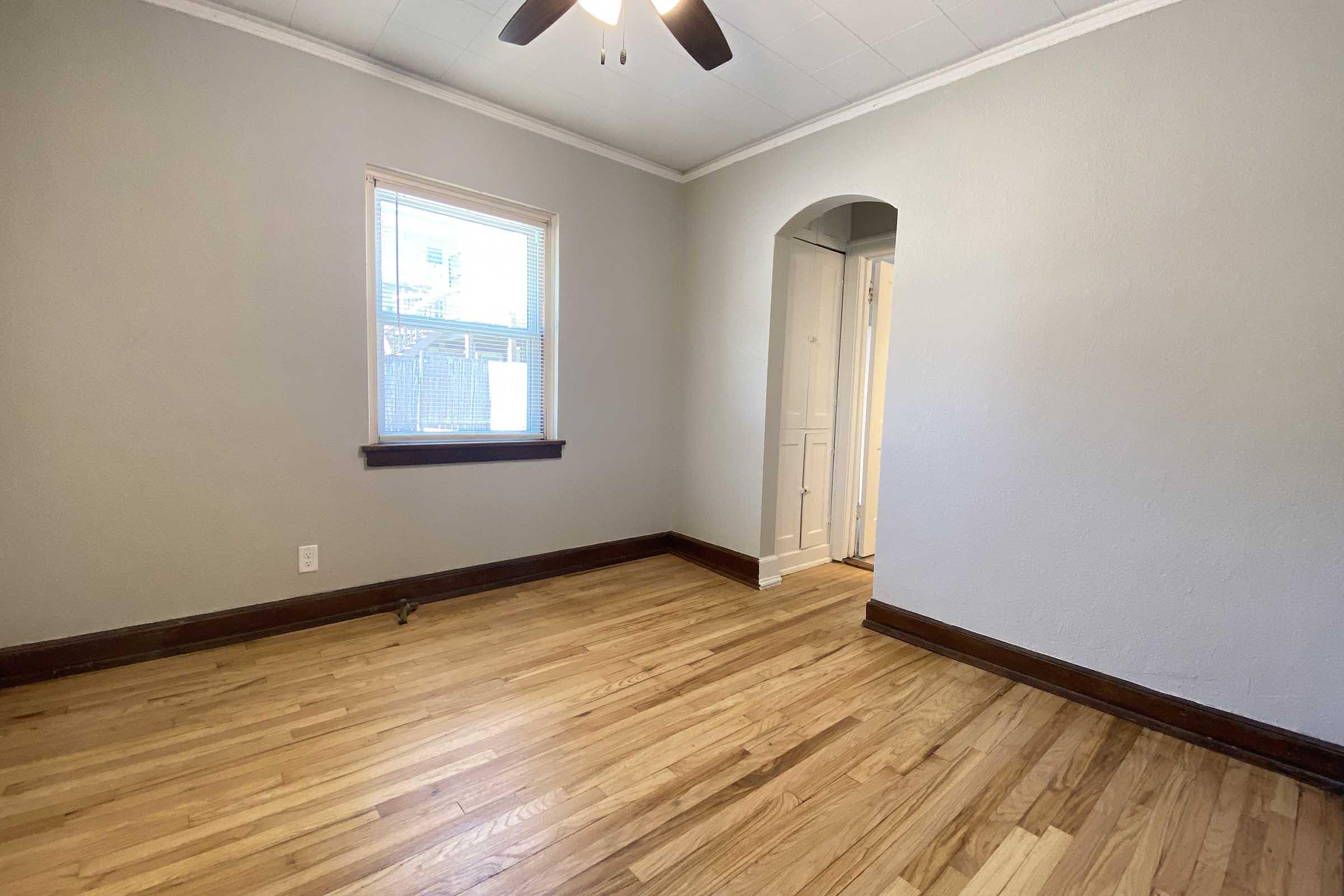 Empty room with light gray walls and polished wooden flooring. A window on the left provides natural light, and a ceiling fan is visible. An archway leads to a hallway on the right, revealing a door. The space has a clean, minimalist feel, suitable for various uses like a bedroom or office.