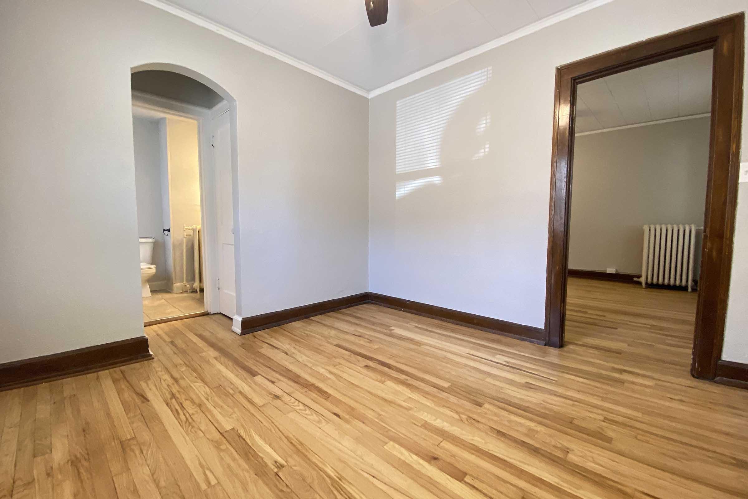 A spacious room with hardwood flooring, light gray walls, and a ceiling fan. An open doorway leads to a bathroom, visible in the background. Another doorway on the right opens into a hallway. Soft natural light brightens the space through a window, highlighting the clean, minimalist design.