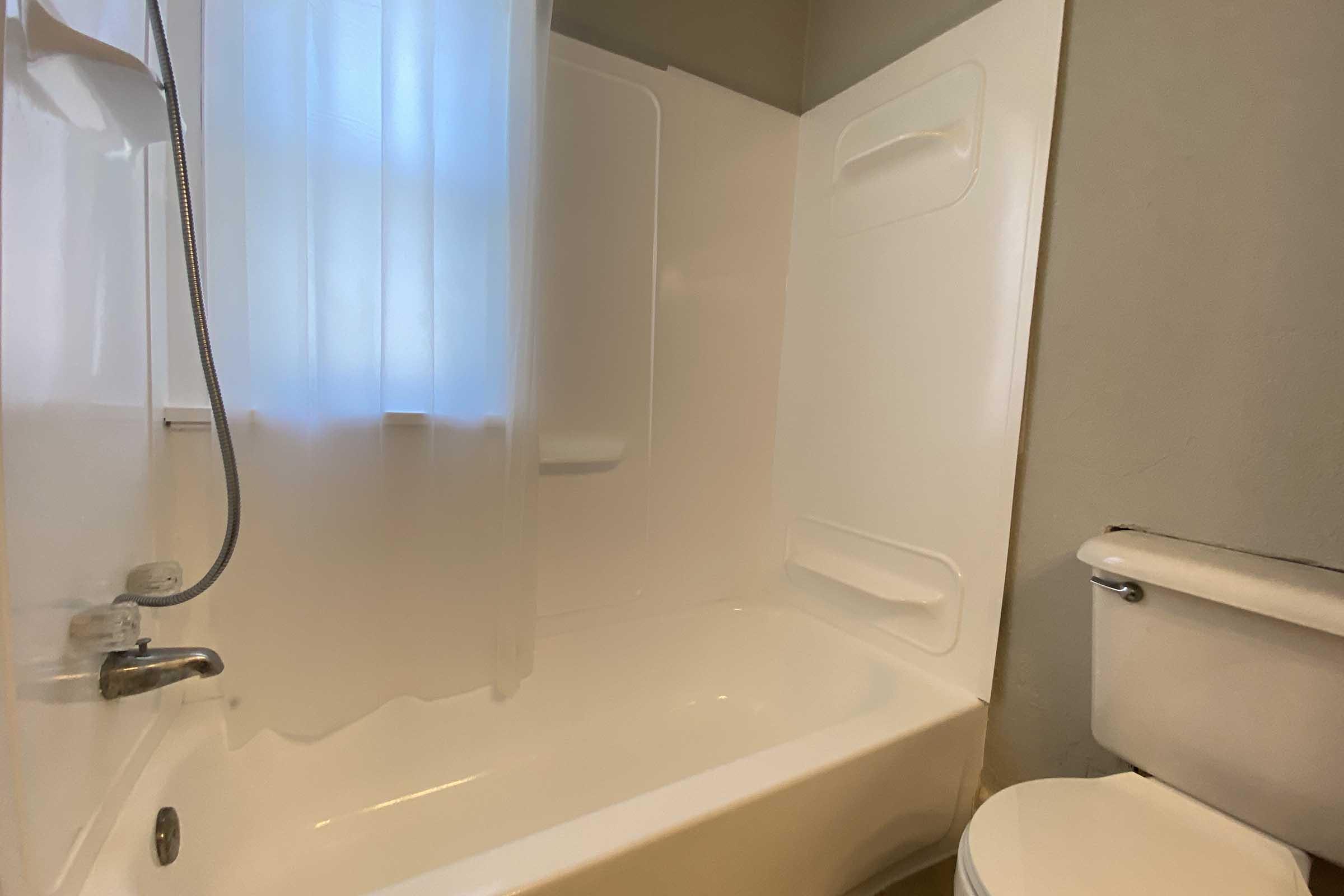 A clean and simple bathroom with a white bathtub and shower, a showerhead on the wall, a light-colored curtain partially covering the window, and a white toilet adjacent to the bathtub, all set against light-colored walls.