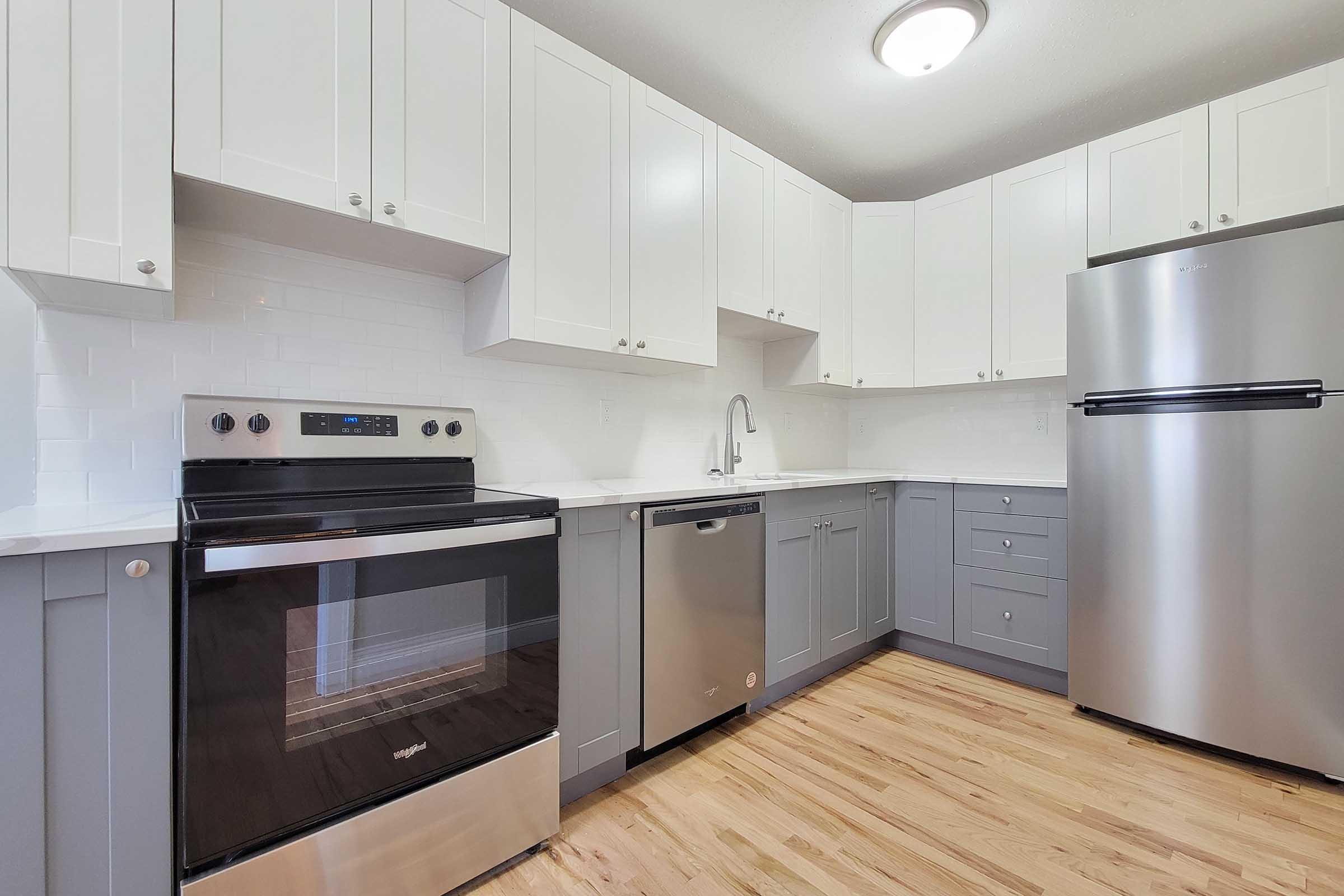 Modern kitchen featuring white and gray cabinetry, stainless steel appliances including a refrigerator and oven, a sink with a modern faucet, and hardwood flooring. The space is bright and clean with ample cabinet storage.