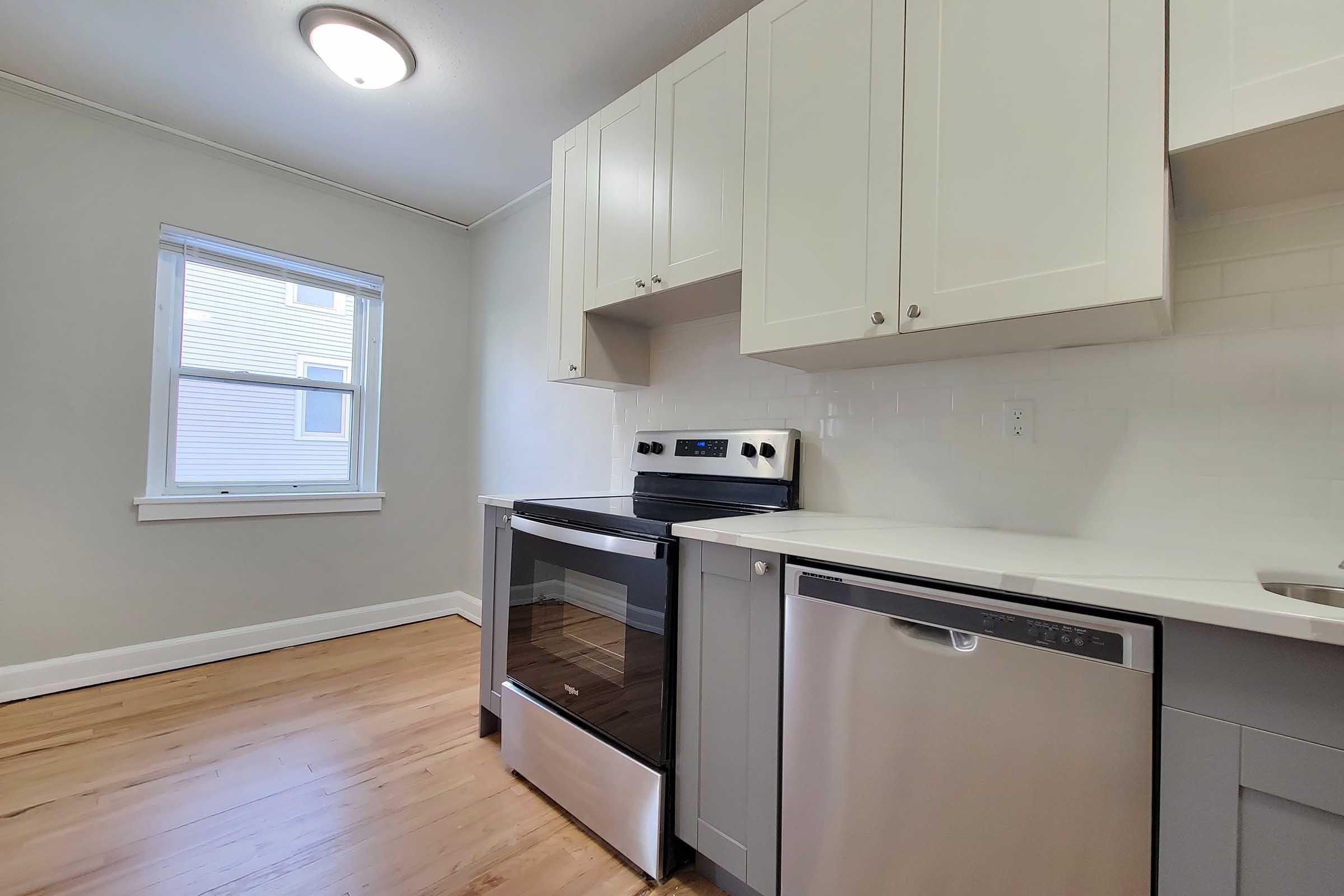 Modern kitchen featuring white cabinetry, a stainless steel oven, and a dishwasher. The room has light-colored flooring and a window that lets in natural light, creating a bright and inviting atmosphere.