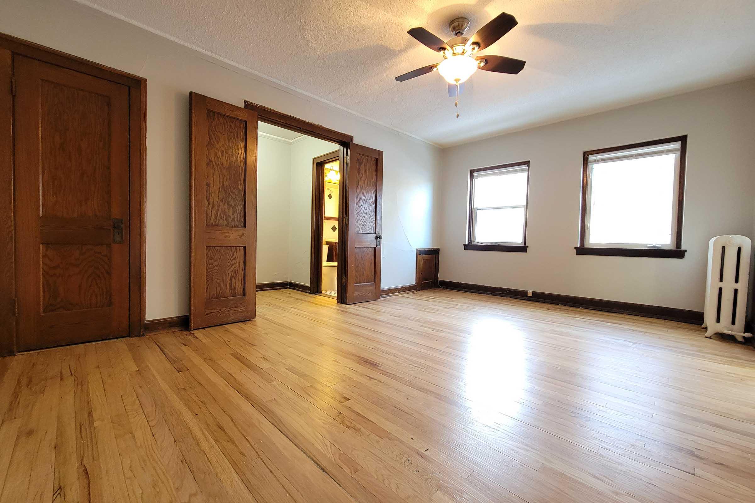 A spacious room with hardwood floors, featuring two wooden doors on the left, three windows allowing natural light, and a ceiling fan. A bathroom door is visible in the background. The walls are painted in a neutral color, adding to the bright and open feel of the space.