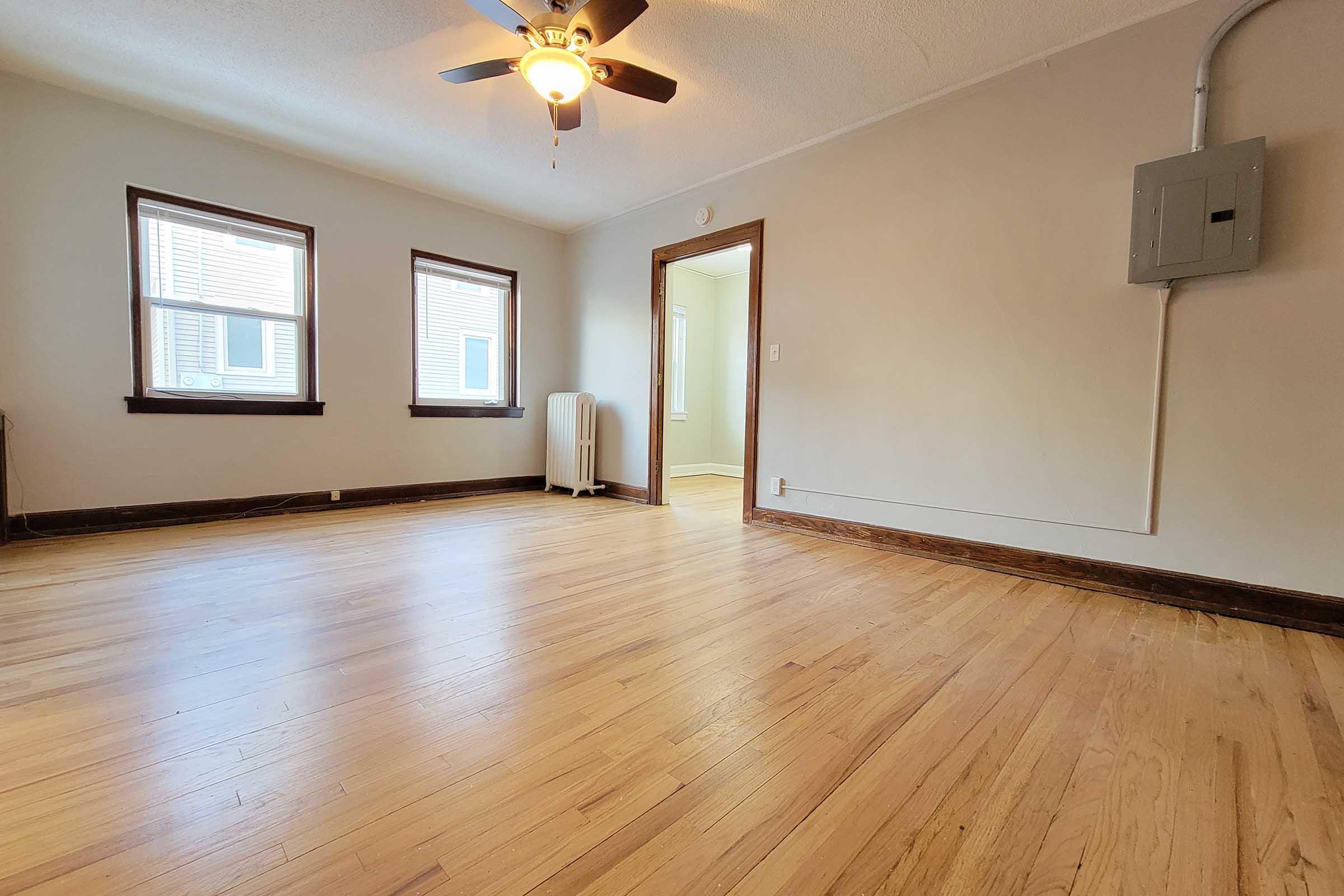 A spacious room with hardwood floors, featuring three windows allowing natural light. There is a ceiling fan for airflow and a doorway leading to another area, possibly a hallway or additional room. A radiator is visible along one wall, adding to the room's vintage charm.