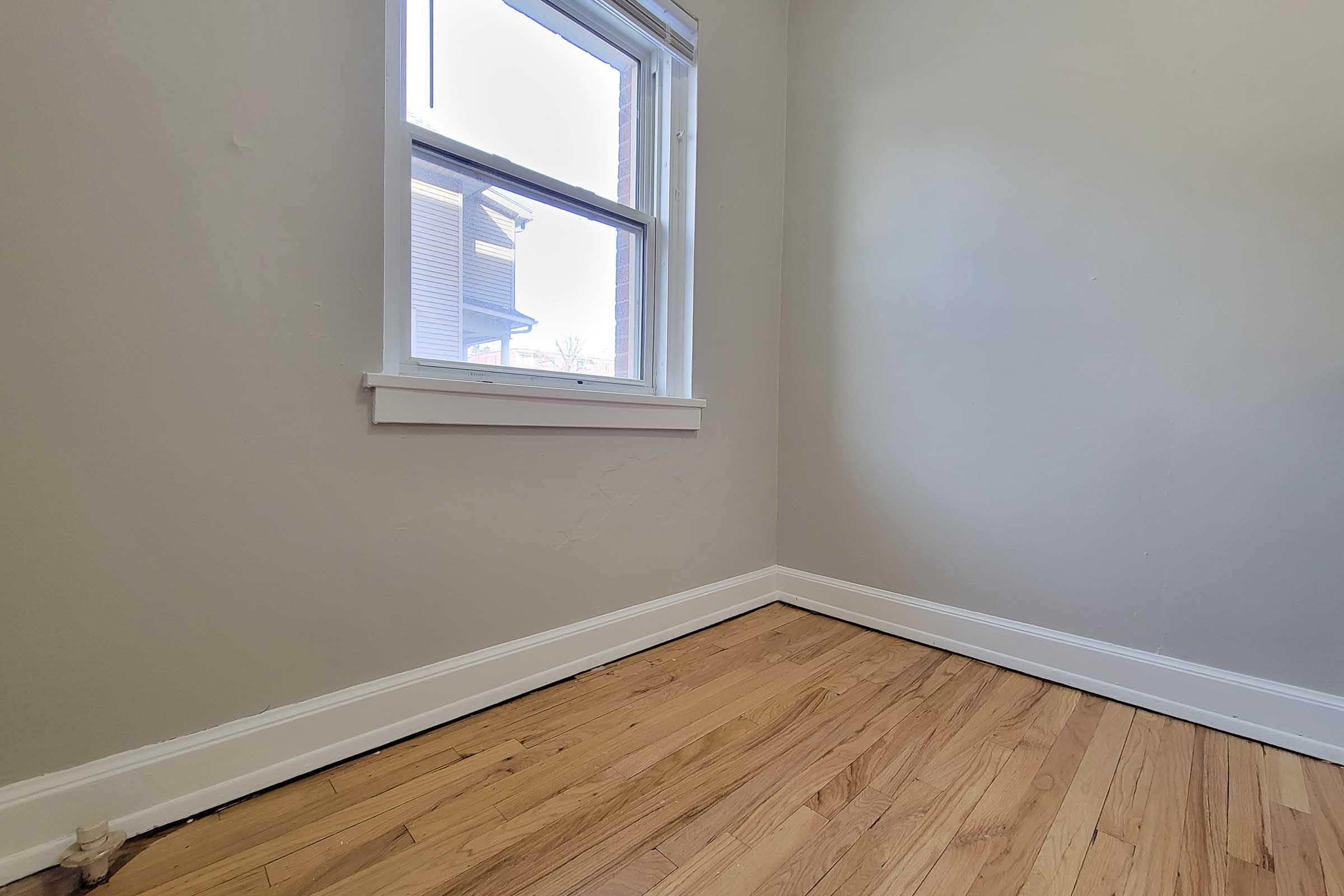 A corner of a room featuring a window that lets in natural light. The walls are painted in a light color, and the floor has wooden planks. The space is empty, giving it a clean and minimalist look.