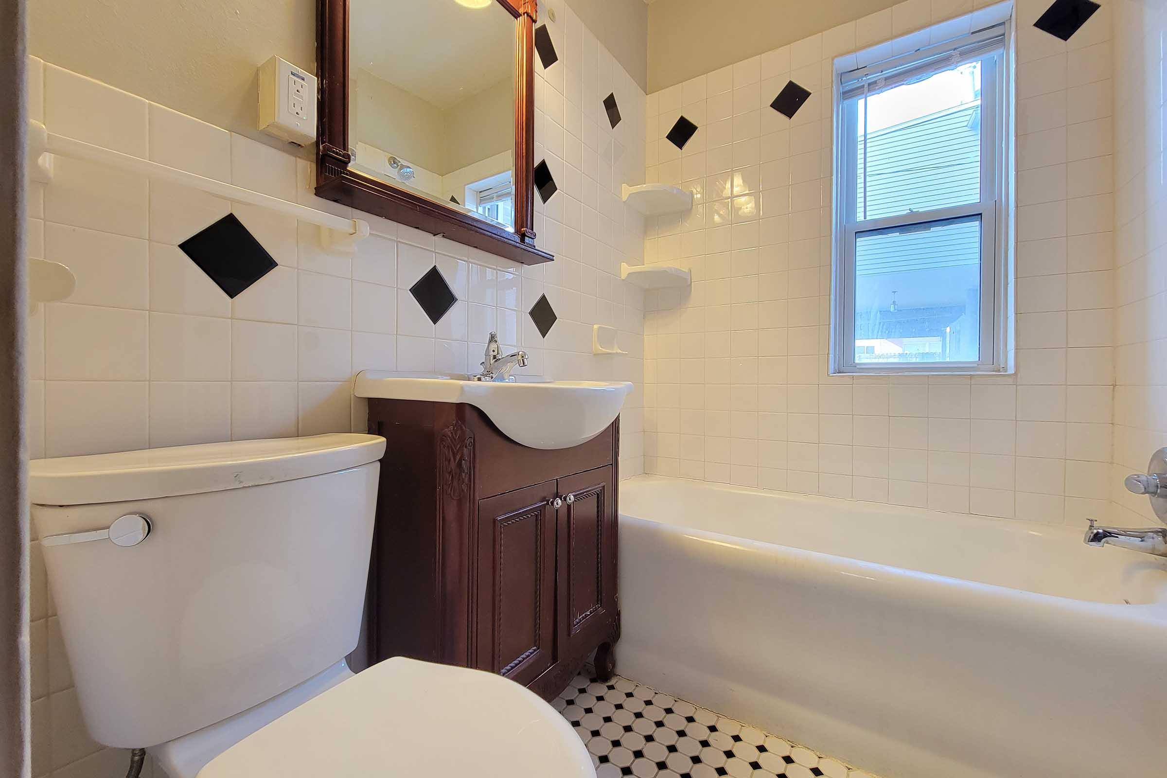 A clean, well-lit bathroom featuring a white bathtub, a toilet, and a wooden sink vanity. The walls are tiled with white square tiles and black diamond accents. A mirror is above the sink, and there is a small window allowing natural light in. The floor is covered in black and white patterned tiles.