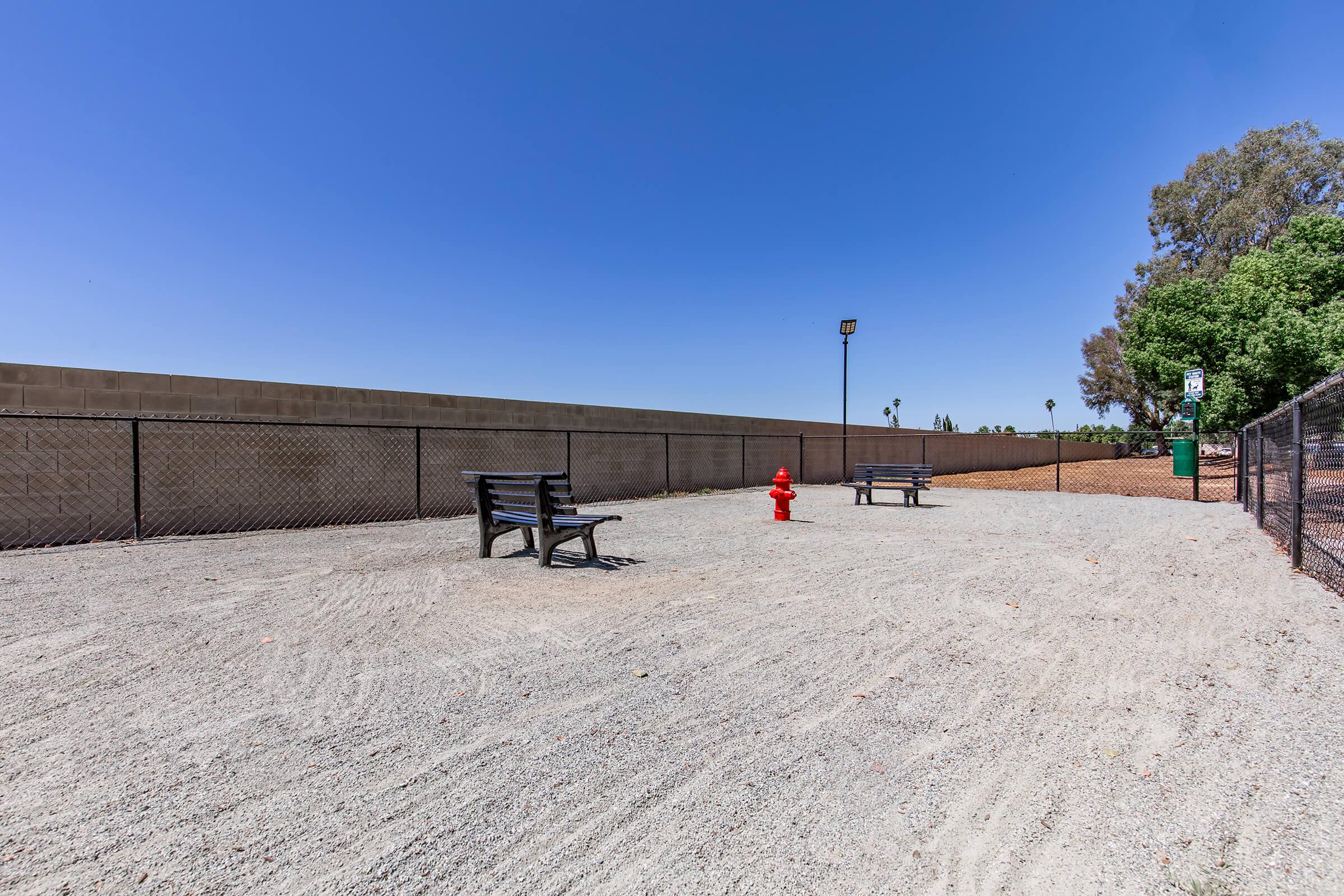 An empty outdoor area featuring two benches on gravel ground, with a red fire hydrant situated nearby. A chain-link fence encloses the space, and a low wall is visible in the background. The sky is clear and blue, indicating a sunny day.