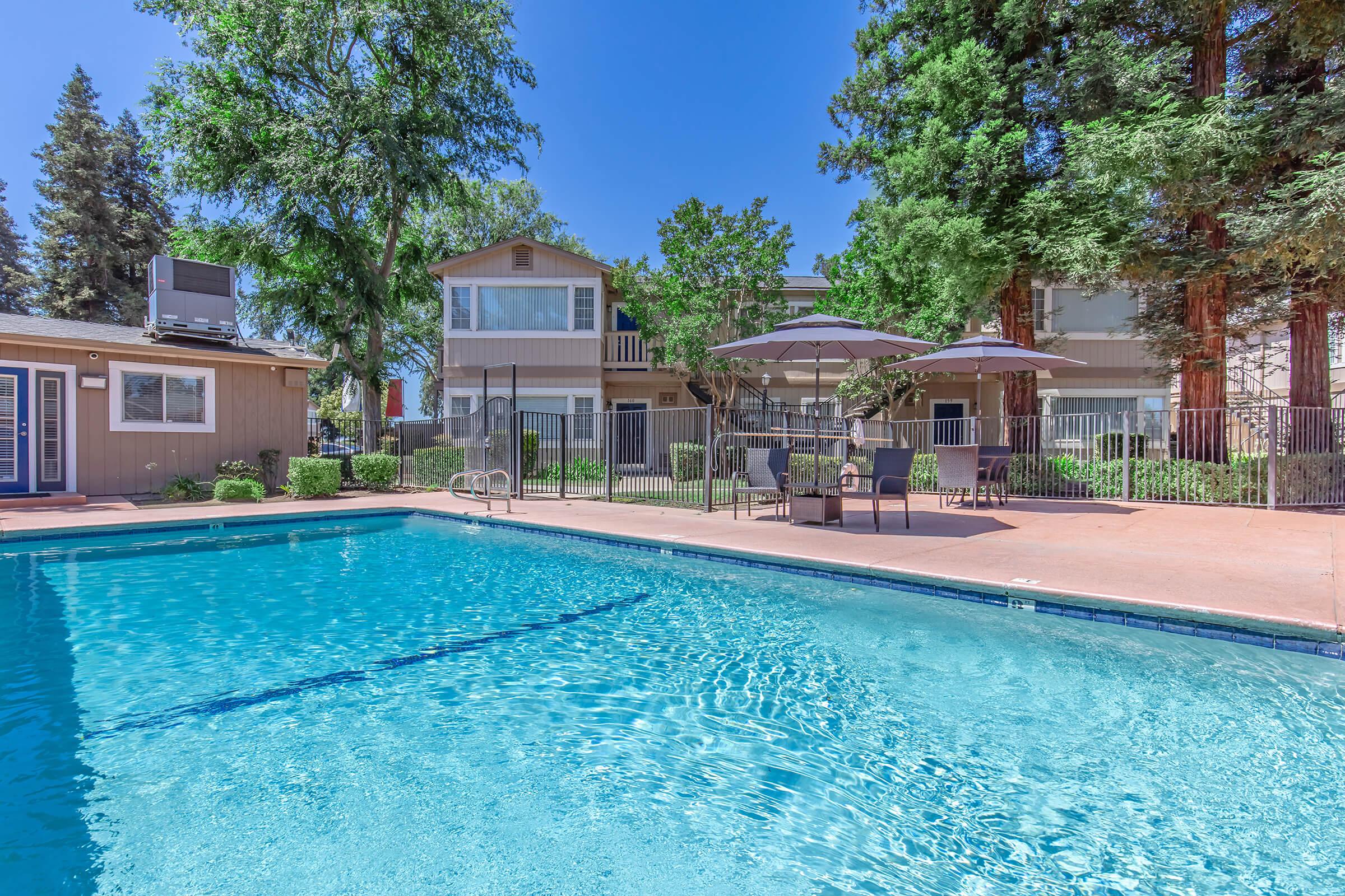 A clear swimming pool surrounded by a patio with lounge chairs and tables. In the background, there are trees and a residential building. The sky is blue and sunny, creating a relaxing outdoor atmosphere.