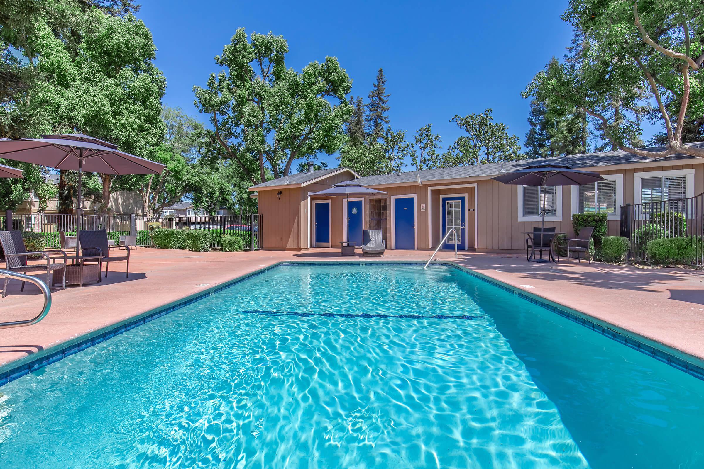 A sparkling blue swimming pool surrounded by a patio area with lounge chairs and umbrellas. In the background, there are several buildings with blue doors, lush greenery, and trees providing shade, creating a relaxed outdoor atmosphere. The sky is clear and blue, enhancing the inviting ambiance.