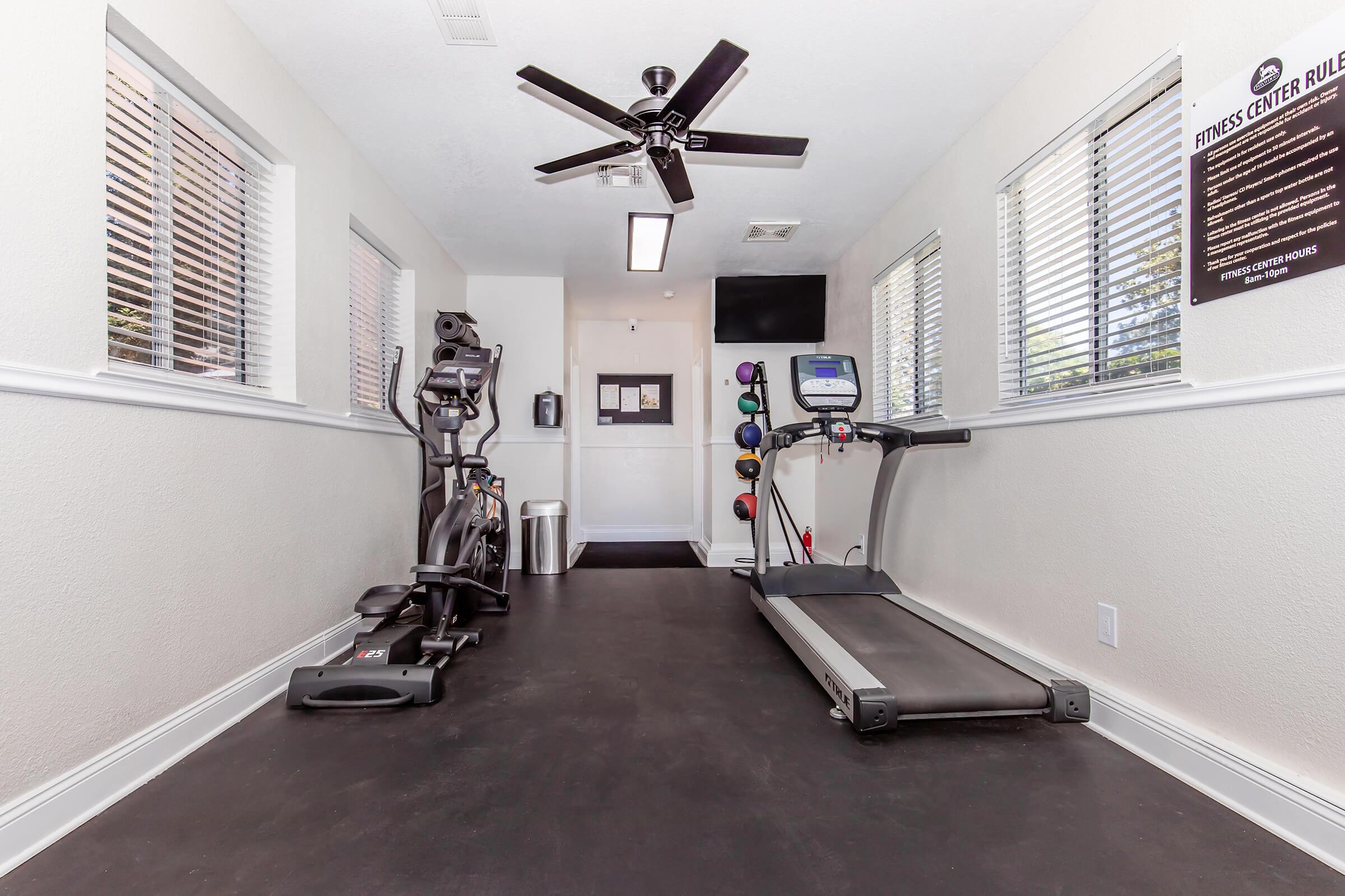 A small fitness center featuring an elliptical machine, a treadmill, and a wall-mounted set of dumbbells. The room has large windows for natural light, a ceiling fan, and a printed poster outlining fitness center rules. The flooring is dark and the walls are painted a light color.
