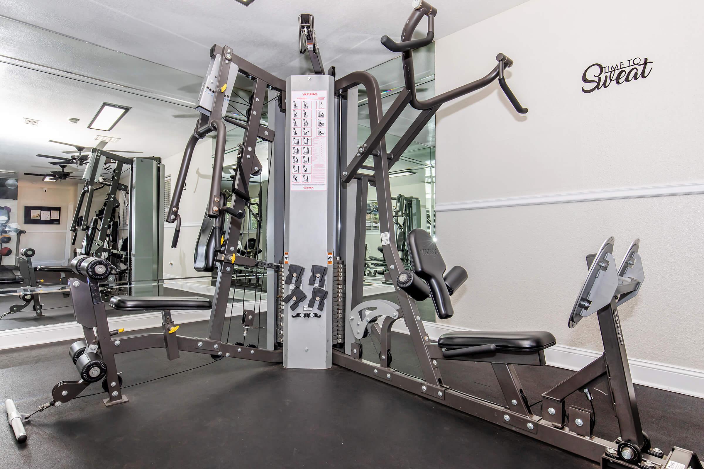 A compact home gym featuring a multifunctional strength training machine with various exercise stations, including a bench press and pull-up bars. The walls are mirrored, and there is motivational text on the wall reading "Time to Sweat." The space is well-lit and designed for fitness activities.