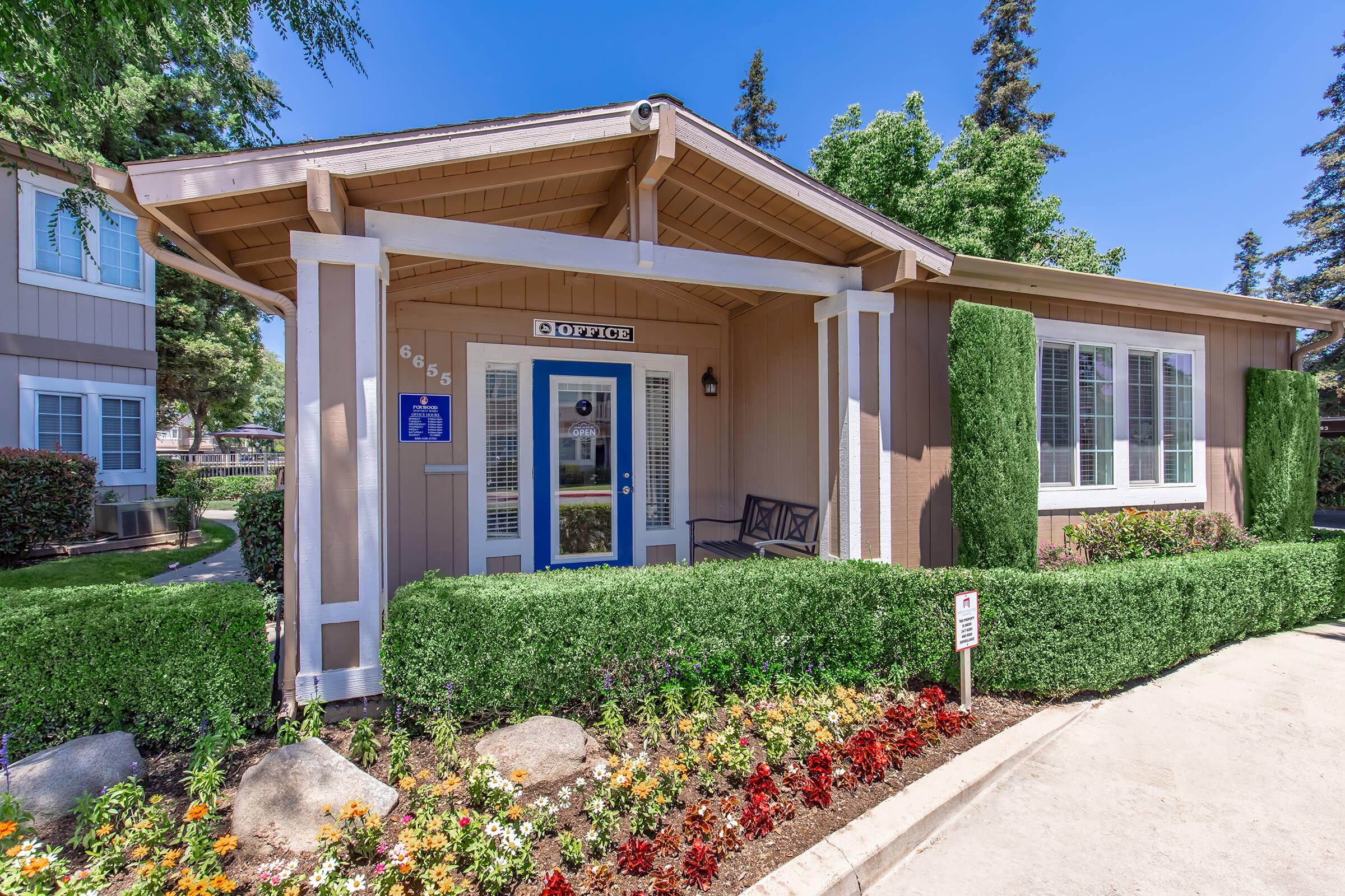 A well-maintained office building surrounded by neatly trimmed hedges and colorful flower beds. The entrance features a blue door and sign labeled "Office," with a bench nearby. Tall trees and clear blue skies complete the inviting outdoor setting.