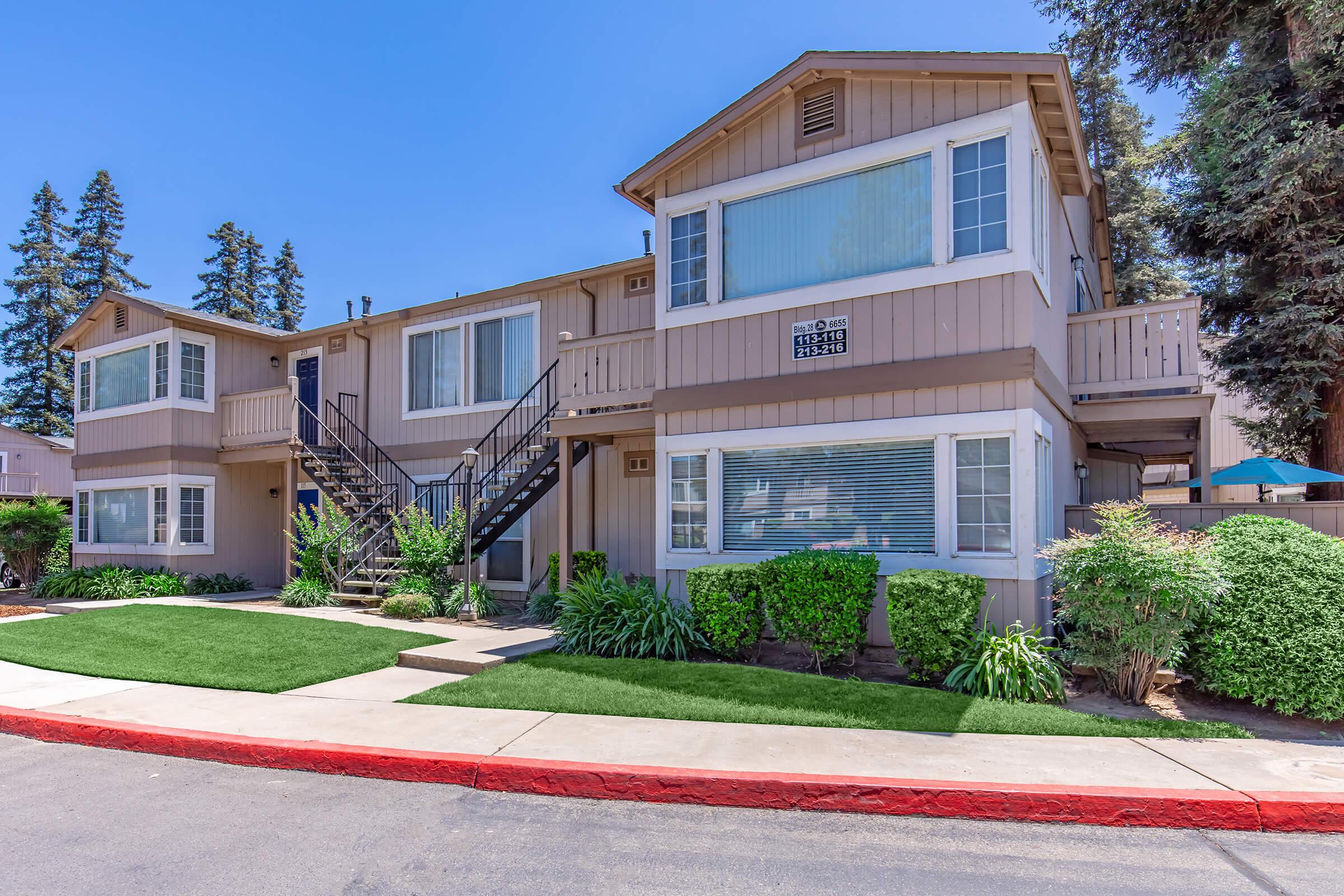 Two-story residential apartment building with light beige siding and dark shutters. Stairs lead to the second floor. The lawn is well-maintained with bushes. Blue sky and tall trees are visible in the background, indicating a clear day. The sidewalk curves around the building.