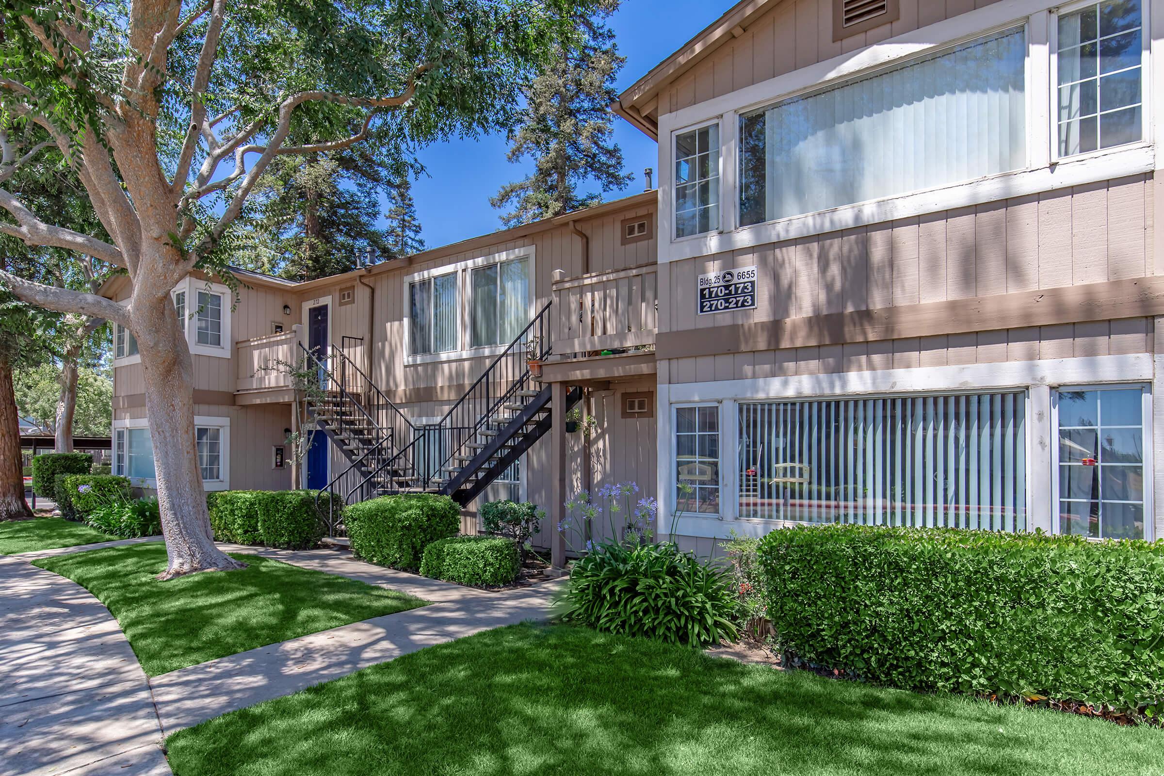 A well-maintained two-story apartment building with beige siding and white trim, featuring a staircase on the left side. Lush green grass and manicured bushes line the walkway leading to the entrance. Tall trees provide shade, and the sky is clear and blue, creating a pleasant atmosphere.
