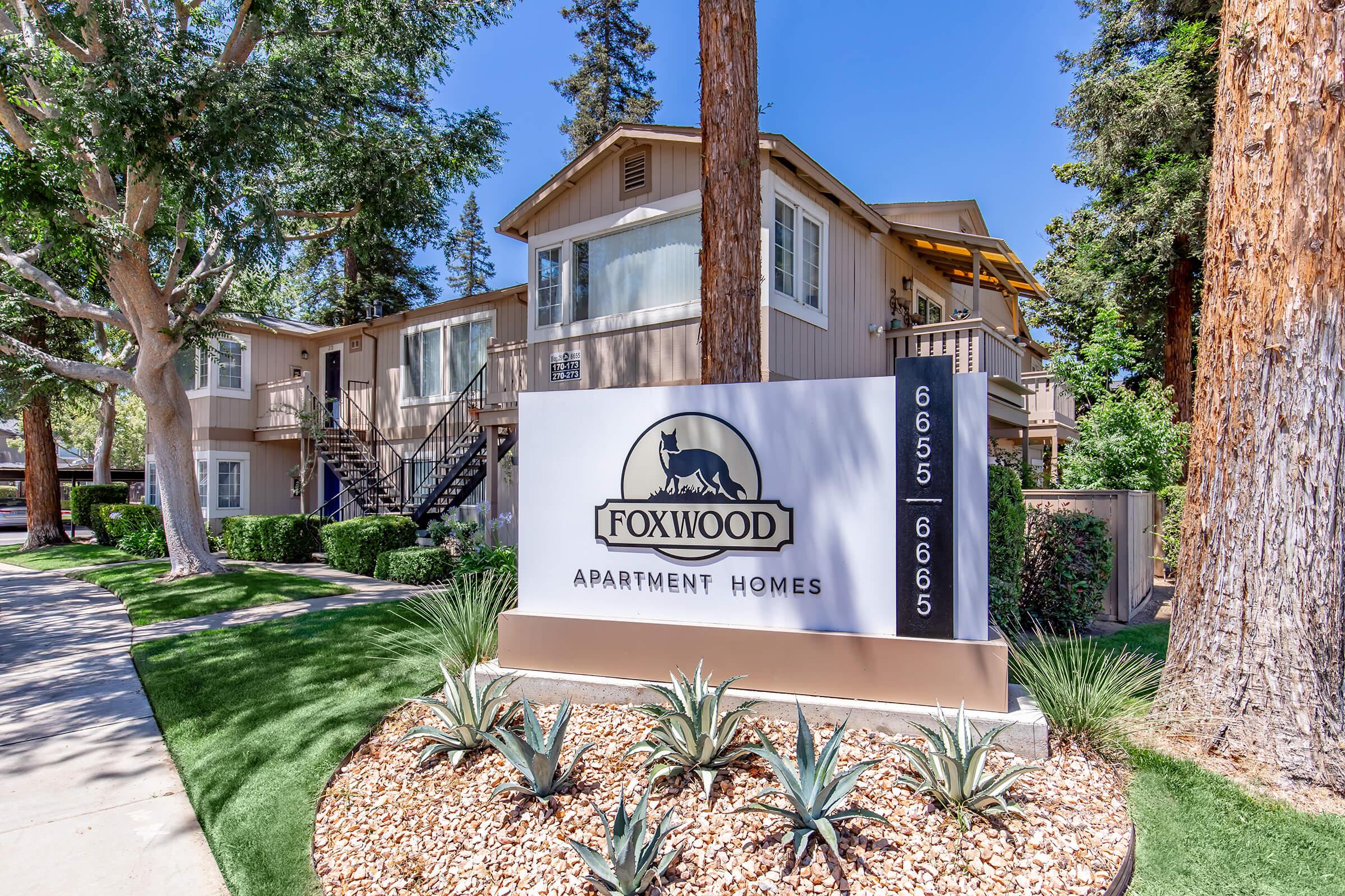 Sign for Foxwood Apartment Homes with the address 6655-6665, surrounded by landscaped greenery, including shrubs and decorative stones. The apartment building features a light brown exterior and large windows, set among tall trees.