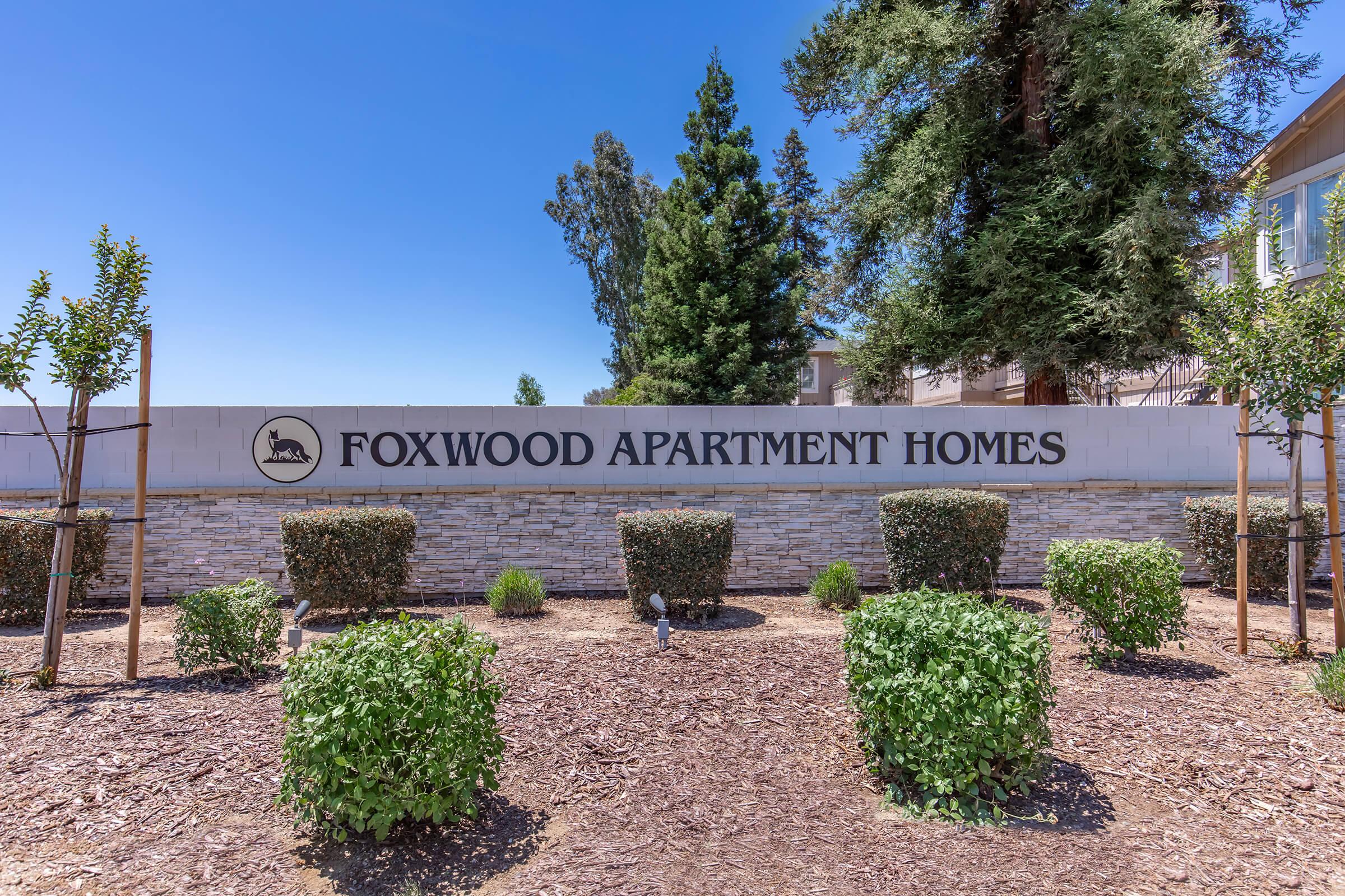 Sign for Foxwood Apartment Homes, featuring neatly trimmed shrubs and trees in a sunny outdoor setting. The sign is prominently displayed against a stone wall, indicating the entrance to the apartment complex. Surrounding landscaping adds a welcoming touch.