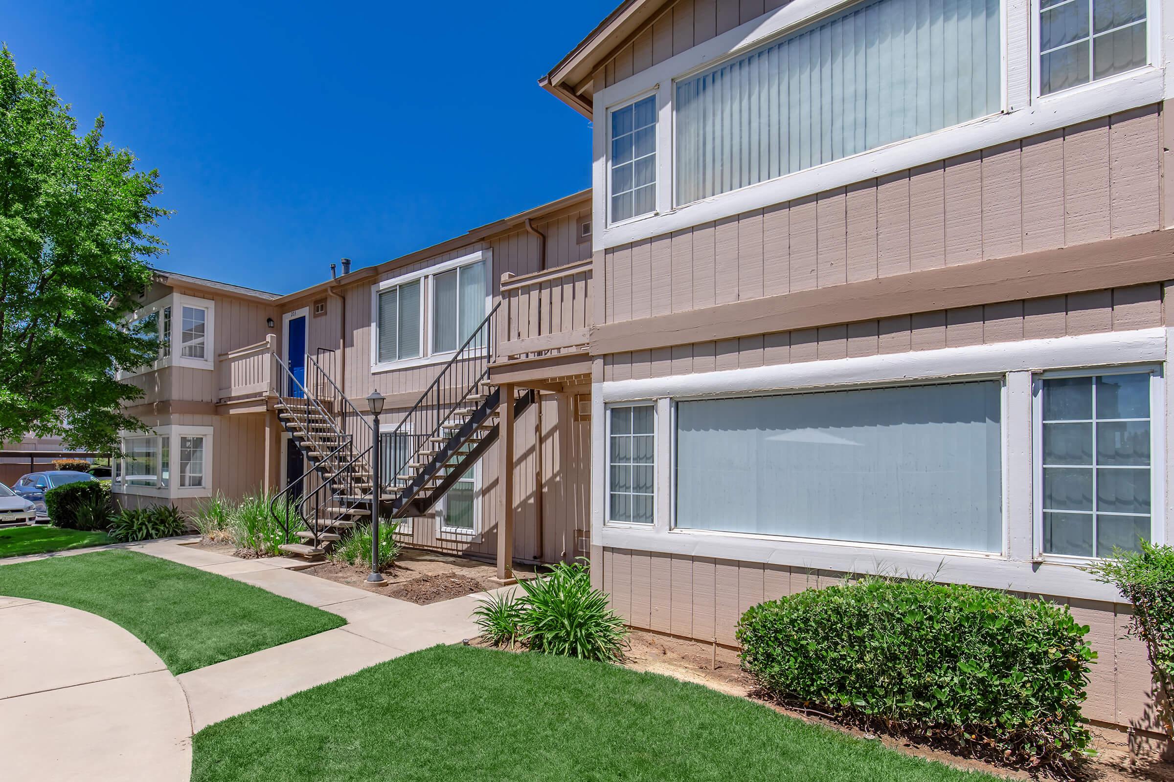 Exterior view of a two-story apartment building with a light brown facade. There are green lawns and bushes in the foreground. A set of metal stairs leads to the upper level. The sky is clear and blue, adding to the bright atmosphere of the scene.
