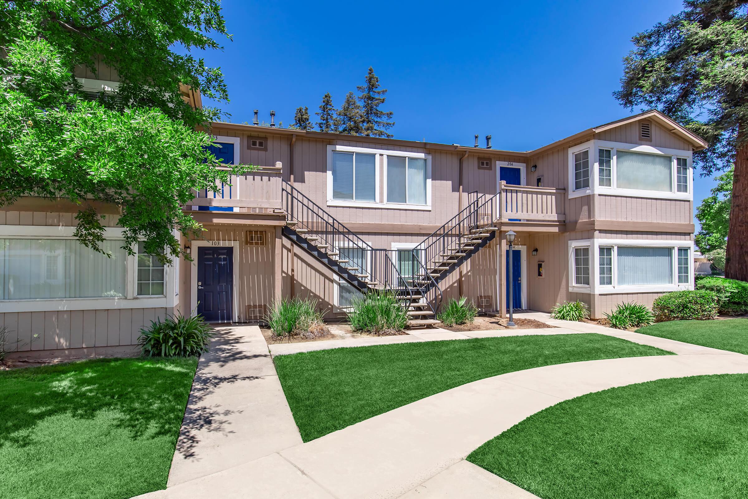 Two-story apartment building with a light brown exterior, featuring two staircases leading to upper-level units. The landscaping includes green grass, shrubs, and trees under a clear blue sky. A concrete pathway curves around the front of the building.