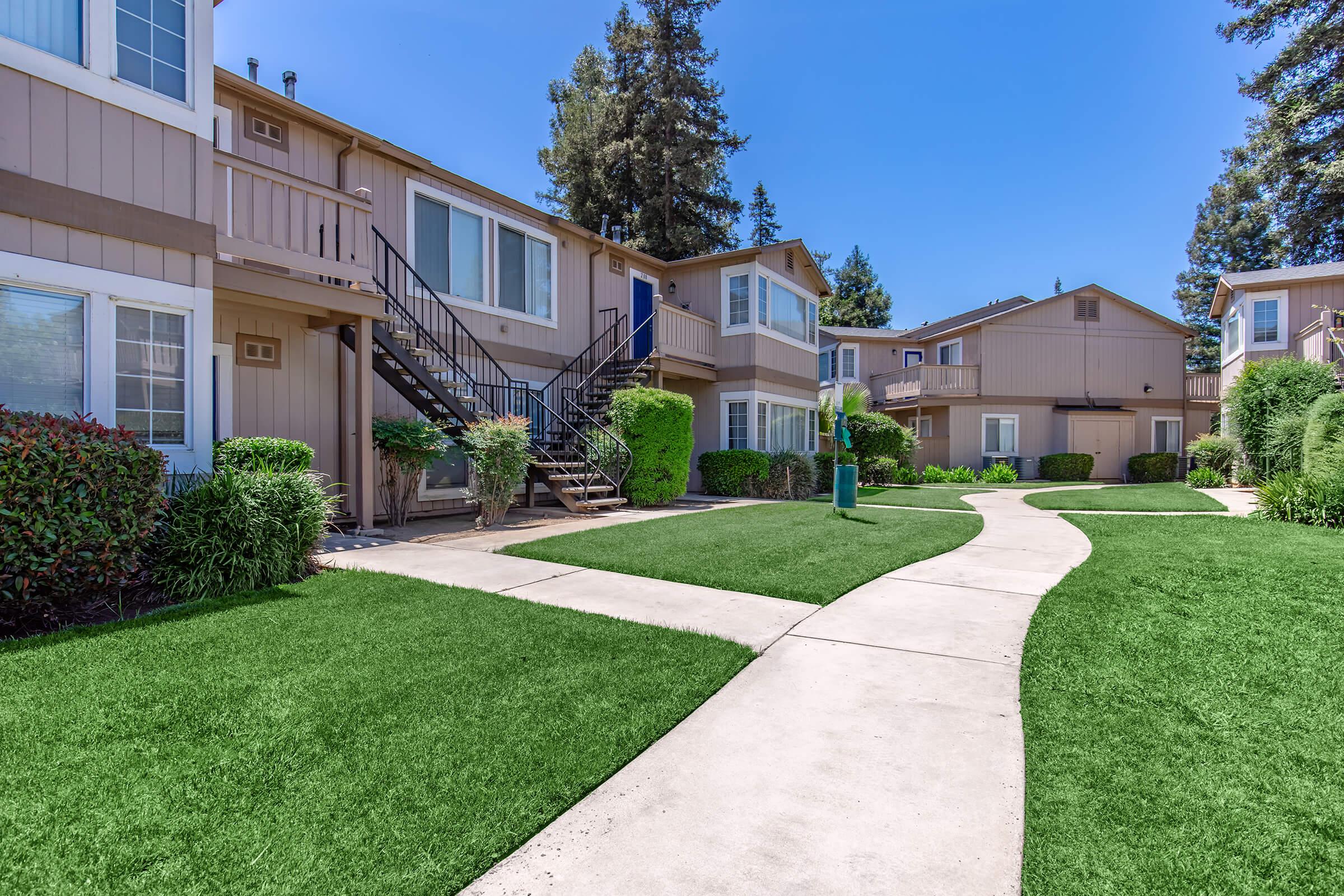 A sunny residential area featuring two-story apartment buildings with light brown exteriors. There is a landscaped lawn with green grass, well-maintained shrubs, and a paved walkway that curves between the buildings, leading to a central green space. Trees and blue skies enhance the inviting atmosphere.