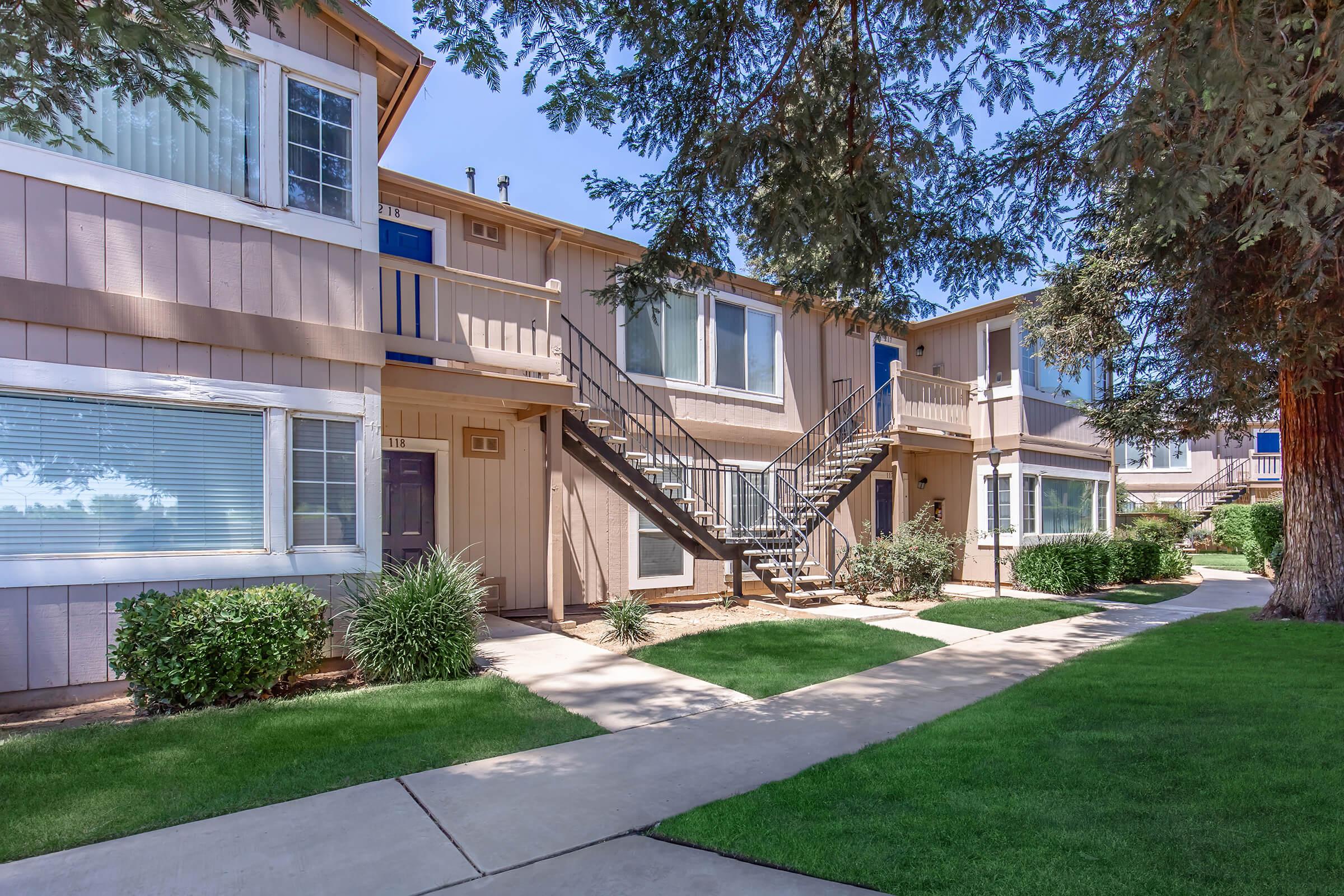 A view of a residential apartment complex featuring two-story buildings with light brown siding. There are balconies and an exterior staircase. Surrounded by well-maintained lawns and shrubs, a paved walkway leads through the property, and it’s a sunny day with clear skies.