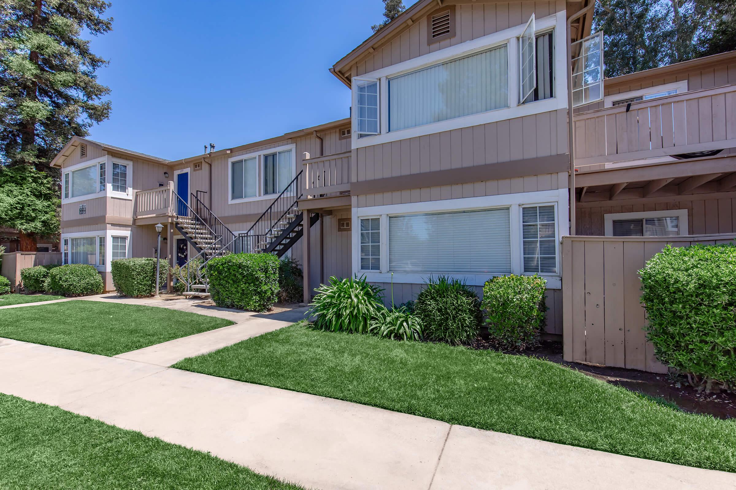 A well-maintained residential building with two stories, featuring beige siding and multiple windows. Lush green bushes and grass line the walkway leading to the entrance, with a staircase on the left side. The scene is set under a clear blue sky, creating a bright and inviting atmosphere.