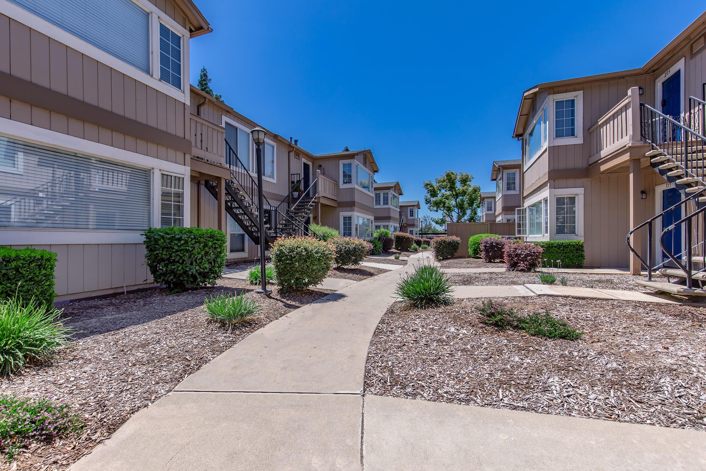 A landscaped pathway leads through a residential complex with two-story apartment buildings. Lush greenery and shrubs line the walkway, creating an inviting atmosphere under a clear blue sky. The buildings feature balconies and staircases, showcasing a well-maintained outdoor space.