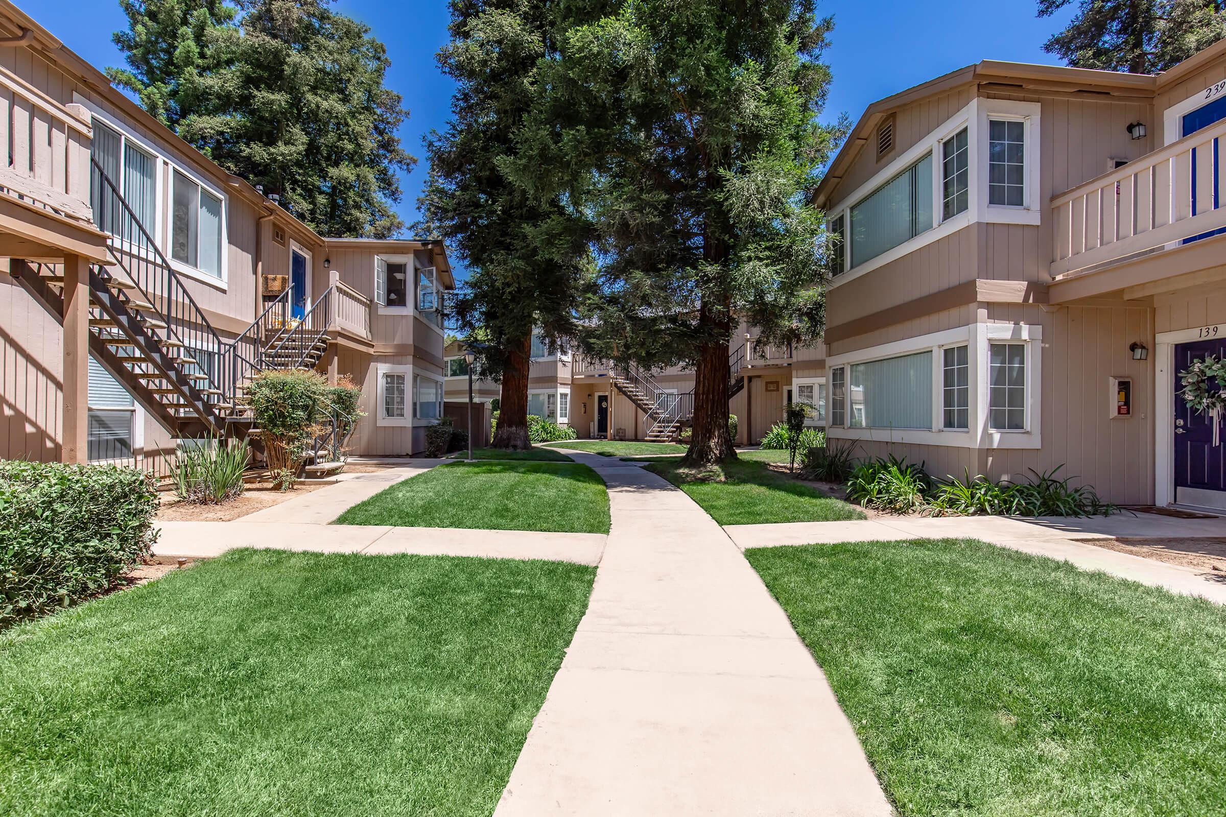 A sunny courtyard featuring two multi-unit buildings on either side. There are green lawns and a paved pathway that splits the landscape, lined with trees. The buildings have balconies and stairs, creating a welcoming, residential atmosphere.