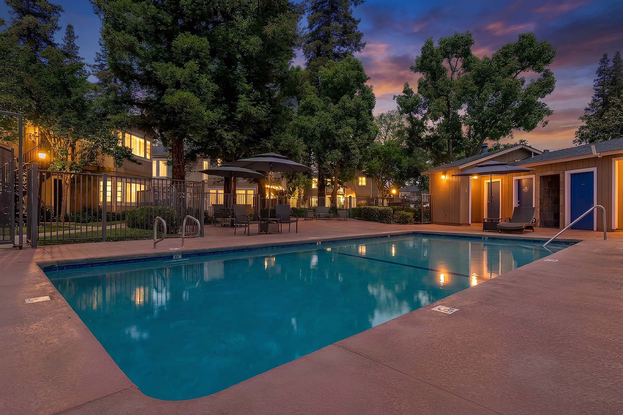A serene swimming pool surrounded by trees and outdoor seating. Soft lighting illuminates the area as the sun sets, casting a warm glow on nearby apartments. The scene is tranquil, featuring a clear blue pool water, creating an inviting atmosphere for relaxation.