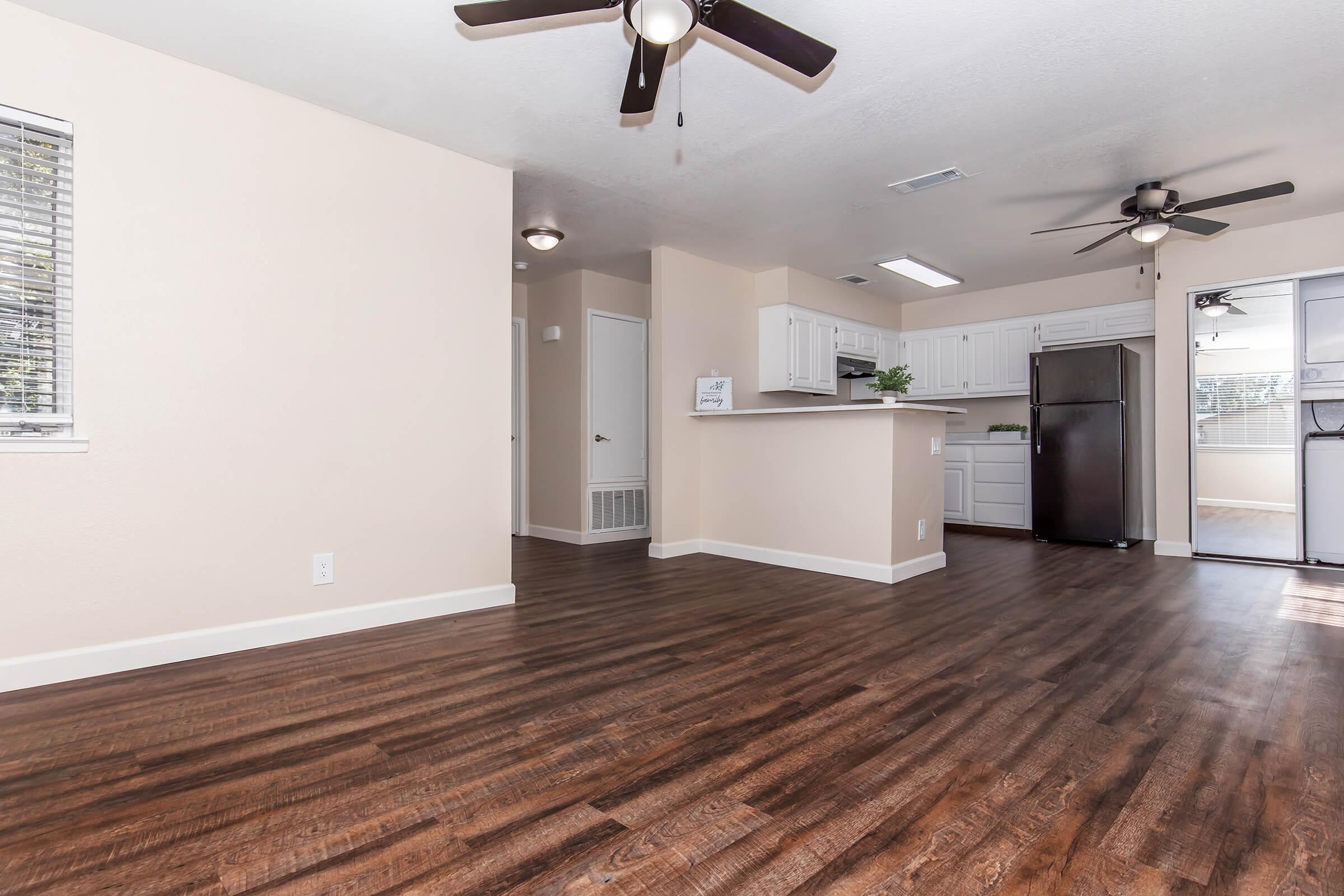 Bright and spacious apartment interior with hardwood-style flooring, featuring a ceiling fan, open kitchen with white cabinetry, and stainless steel appliances. Large windows provide natural light, and there is an entrance to a sunroom or balcony area on the right.