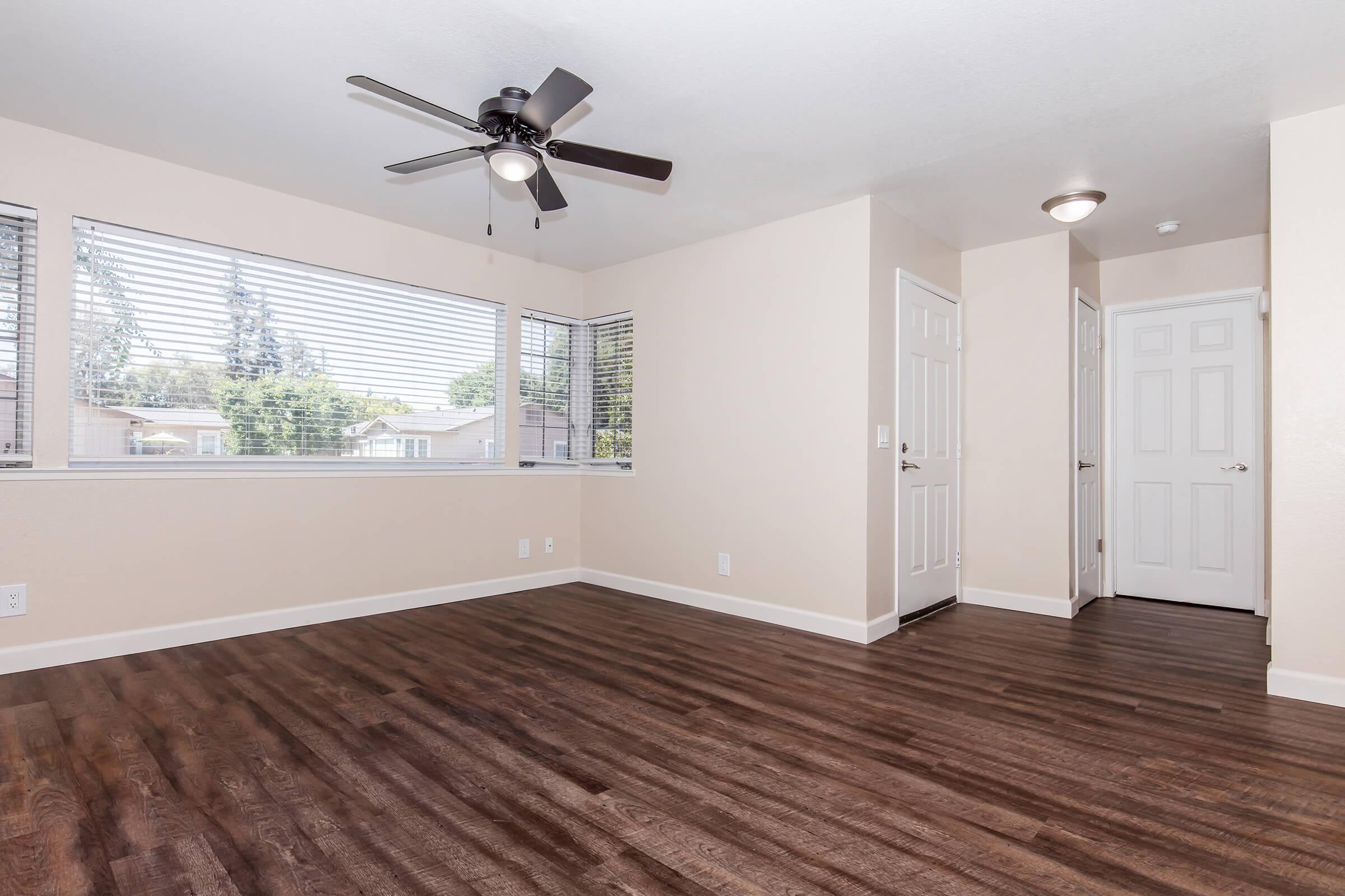 A well-lit, empty room featuring light-colored walls and a ceiling fan. The room has large windows with blinds, showcasing a view outside. There is dark hardwood flooring, and two doors are visible—one leading to a corridor and another possibly to a closet or bathroom.