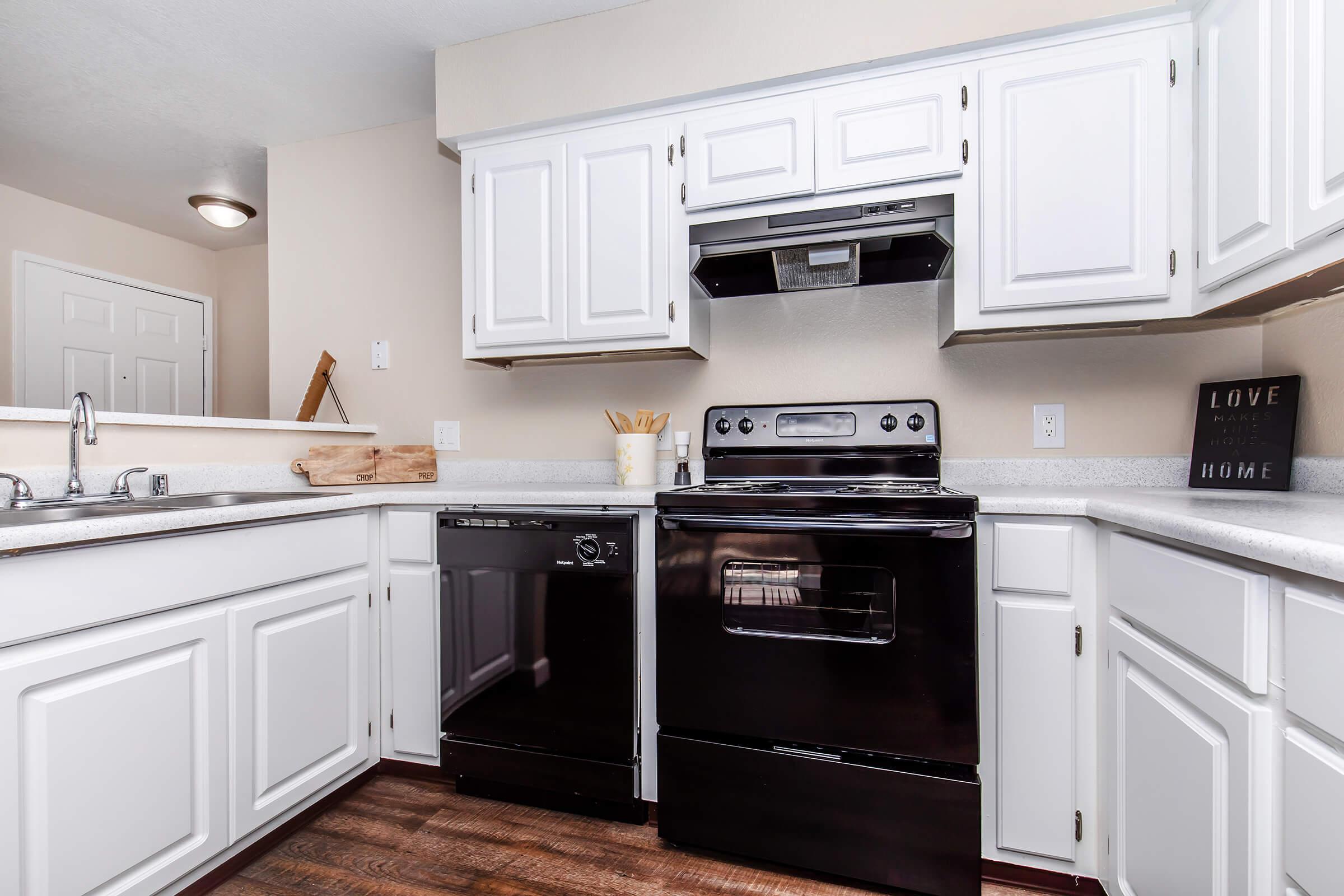 A modern kitchen featuring white cabinets, a black stove, and a black dishwasher. The countertops are light-colored, with kitchen utensils in a container. A wall-mounted range hood is above the stove, and there is a decorative sign on the wall. The flooring is a warm wood tone.