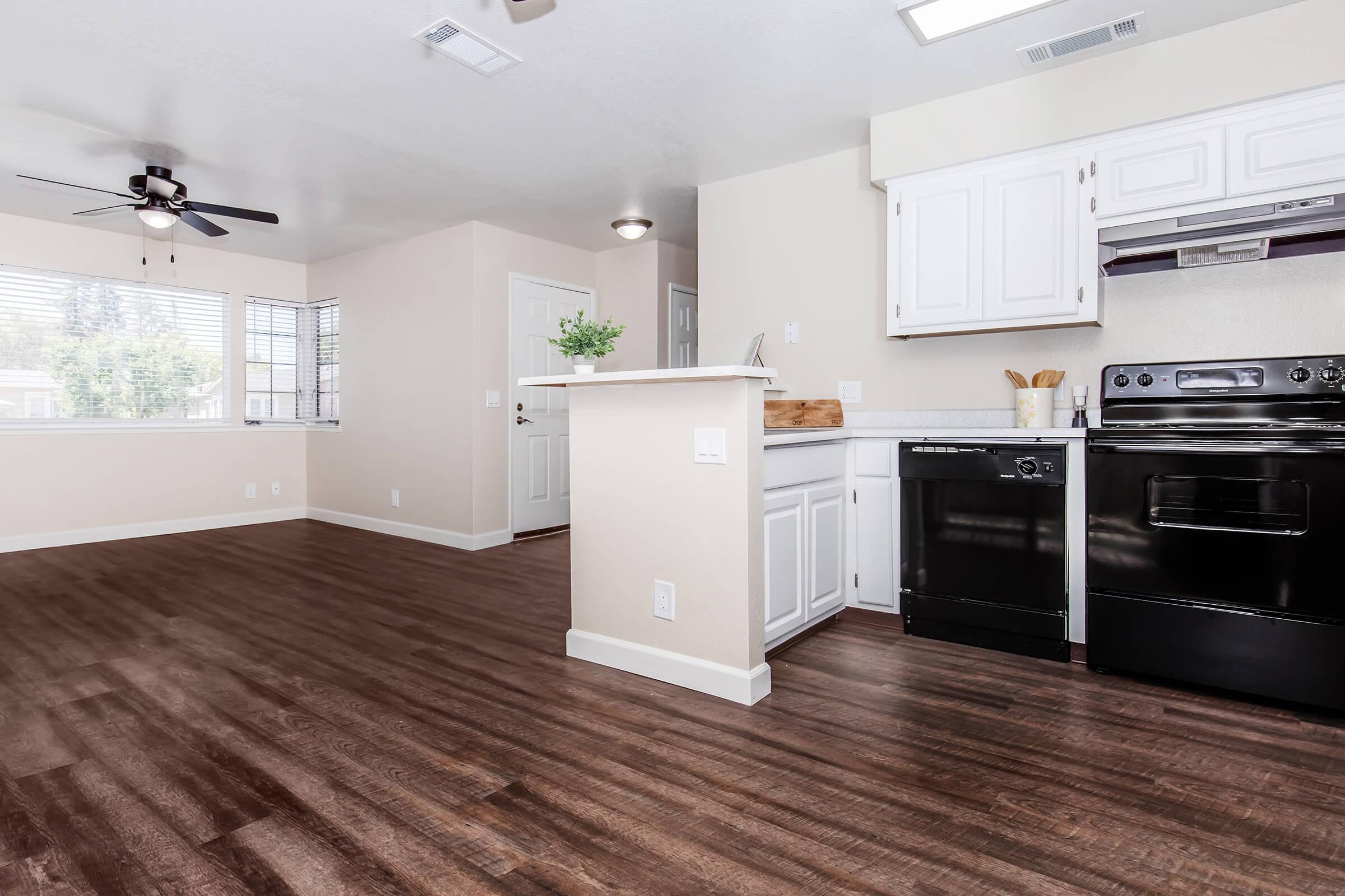 A bright and modern kitchen and living area with dark wood flooring. The kitchen features white cabinets, a black stove, and dishwasher. A ceiling fan is visible and natural light floods in through the windows. The layout is open, connecting the kitchen to the living space, creating a spacious atmosphere.