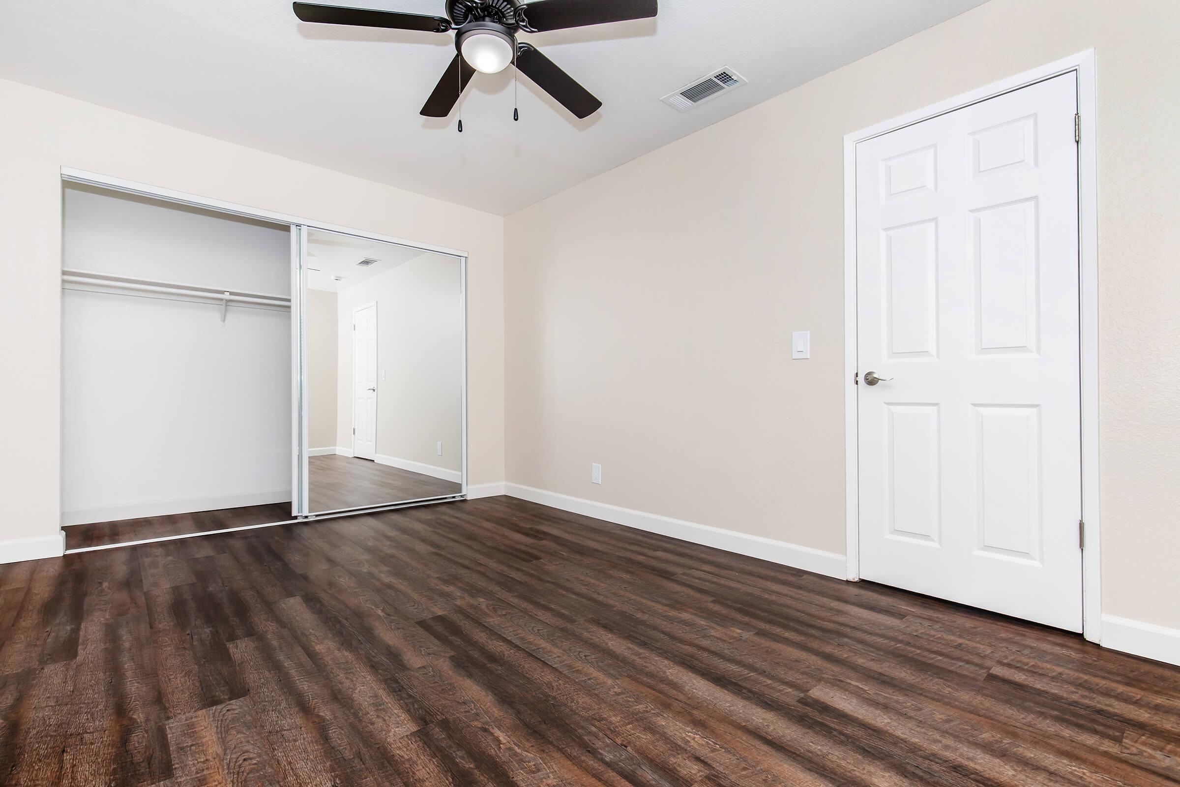 Empty room featuring a wooden floor, light beige walls, a ceiling fan, and a white door. There is a large mirrored closet on one side and another door leading outside the frame. The space is well-lit and appears modern and clean, suitable for various uses.