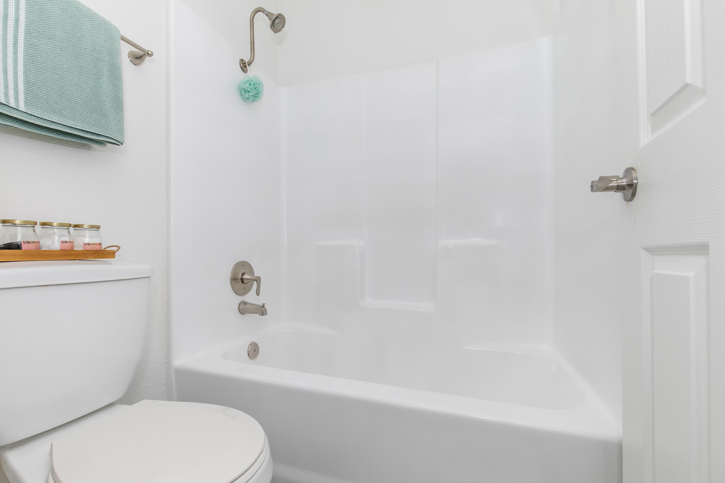 A clean bathroom featuring a white bathtub with attached shower, a toilet, and a small shelf with jars on a wooden surface. Towels are hanging on a rack, and a bath sponge is visible in the shower area, all set against a bright, neutral backdrop.