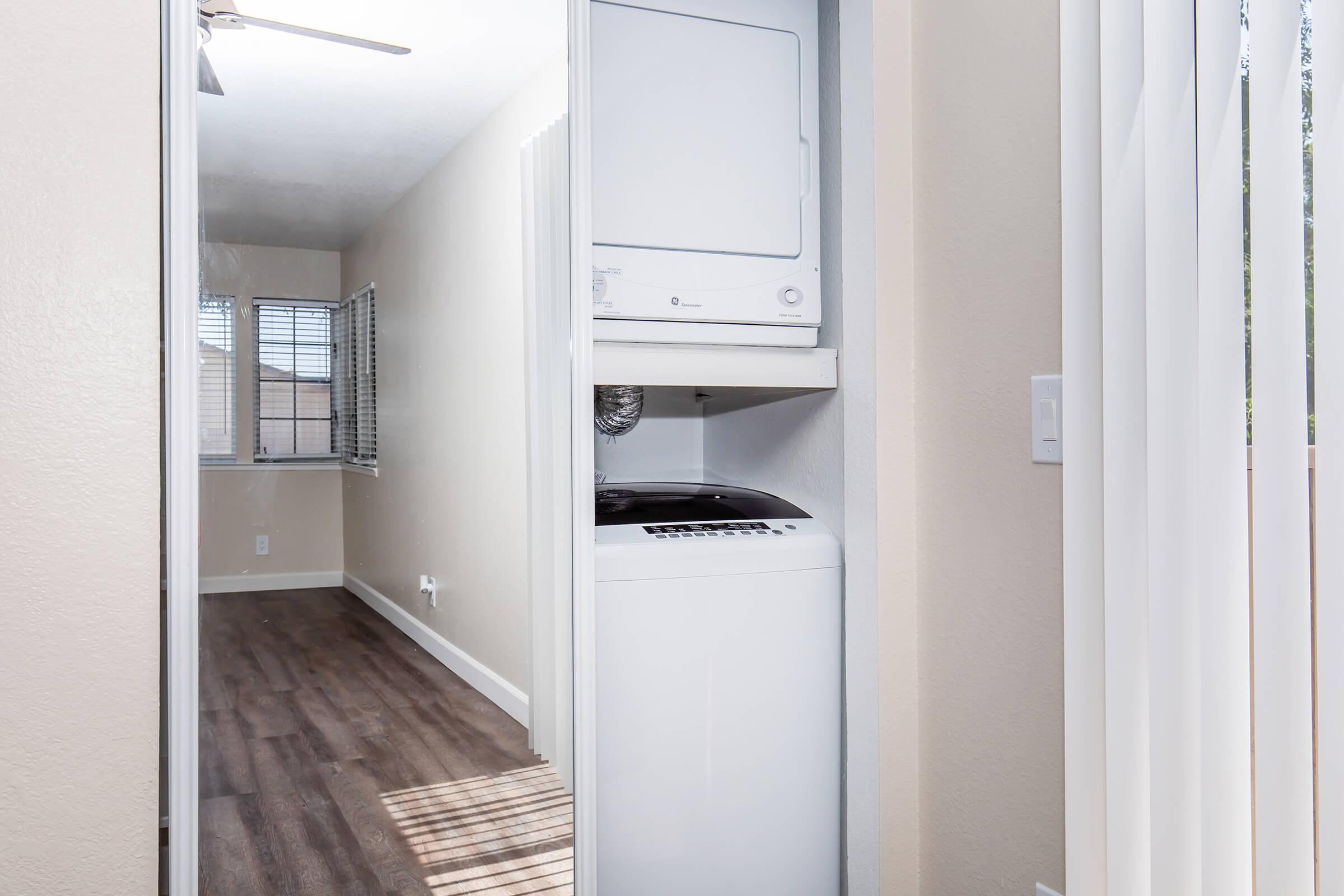 A view of a laundry area featuring a stacked washer and dryer, located in a small room with beige walls and a window. The flooring is made of wood, and the room is well-lit, showcasing a clean and modern design.