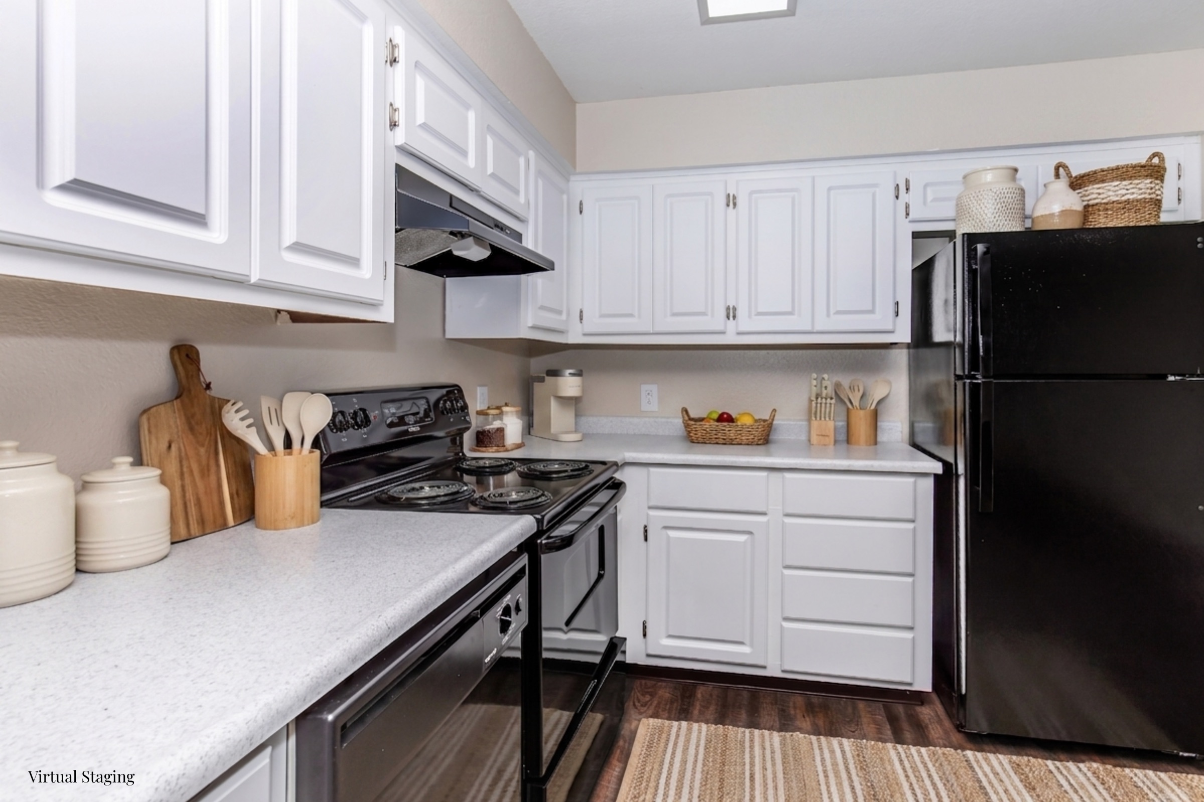 A modern kitchen featuring white cabinets, a black stove and refrigerator, and a light-colored countertop. The kitchen includes wooden utensils, a bowl of fruit, and decorative containers on the counter, with a neutral-toned rug on the floor. The space is bright and well-organized.