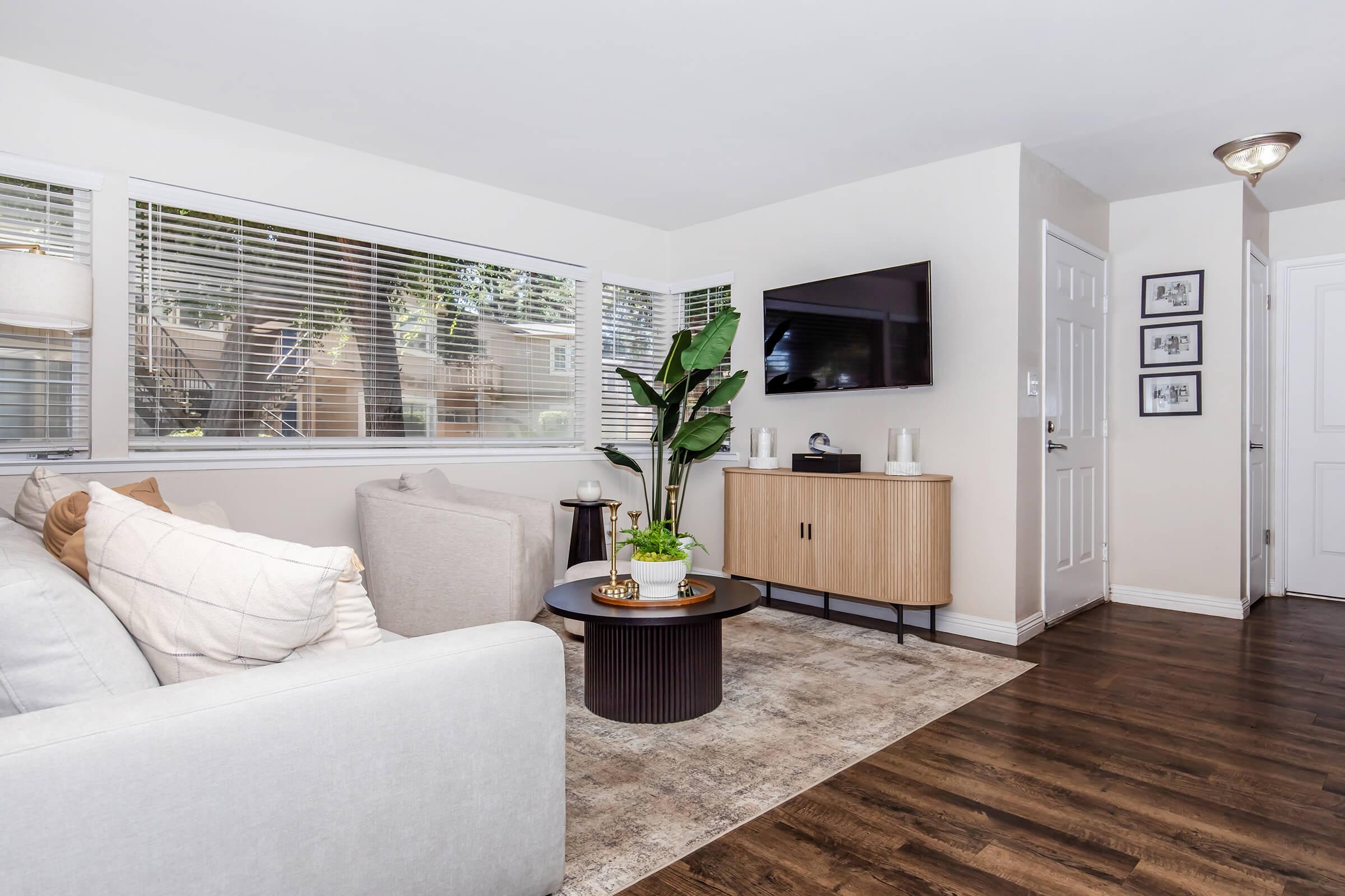 A cozy living room featuring a light-colored couch, a round coffee table with a plant, and a wooden TV stand. Large windows allow natural light to fill the space, complemented by neutral tones and a soft area rug. A door on the right leads to another area of the home.
