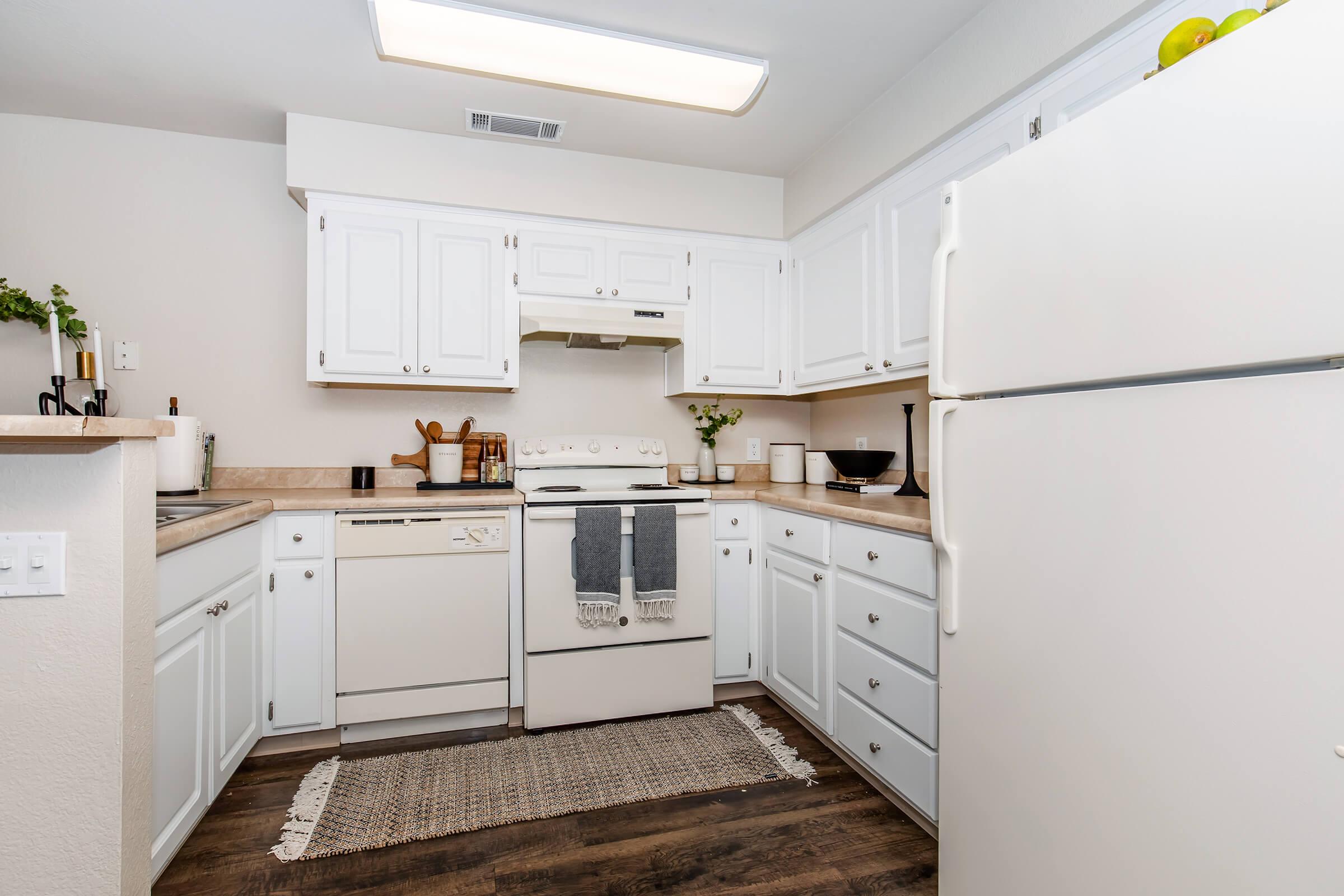 A bright, modern kitchen featuring white cabinetry, a countertop with a small plant, and a rug on the floor. The space includes a white refrigerator, a stove with an oven, a dishwasher, and various kitchen utensils arranged neatly. Natural light fills the room, creating a welcoming atmosphere.