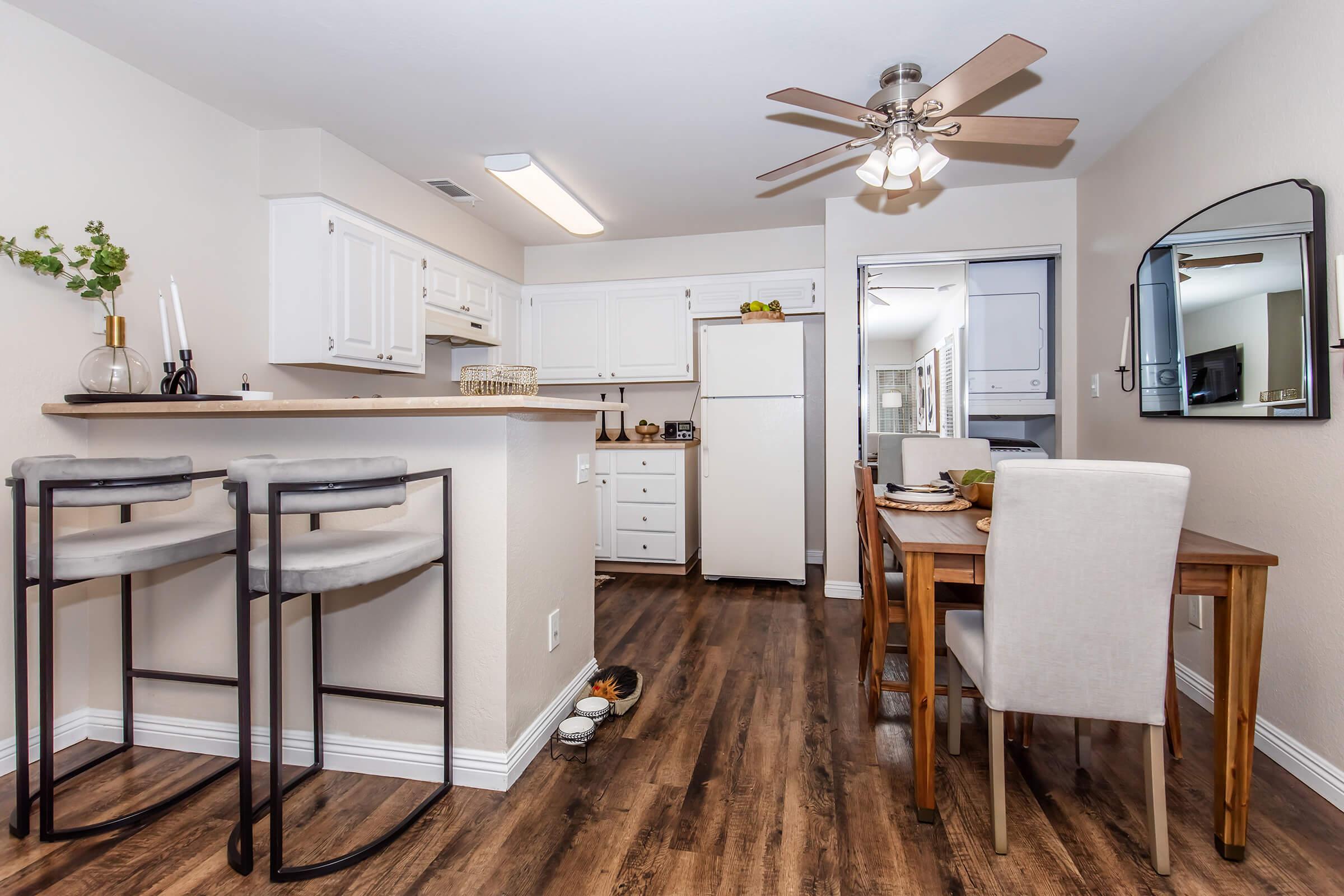 A modern kitchen and dining area featuring white cabinets, a breakfast bar with two stools, a wooden dining table set for two, and a ceiling fan. The flooring is a warm, wooden laminate, and there is natural light coming from a nearby window. Decorative elements include candles and a small plant.