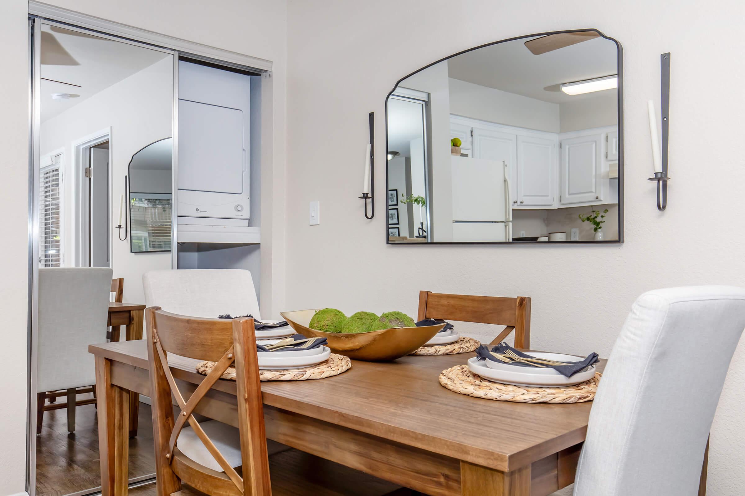 A well-decorated dining area featuring a wooden table set with plates and napkins. A large bowl with decorative items sits in the center. The room is illuminated by natural light, with a mirror reflecting the kitchen area and a washer/dryer unit in the background. Neutral colors and modern accents create a welcoming atmosphere.