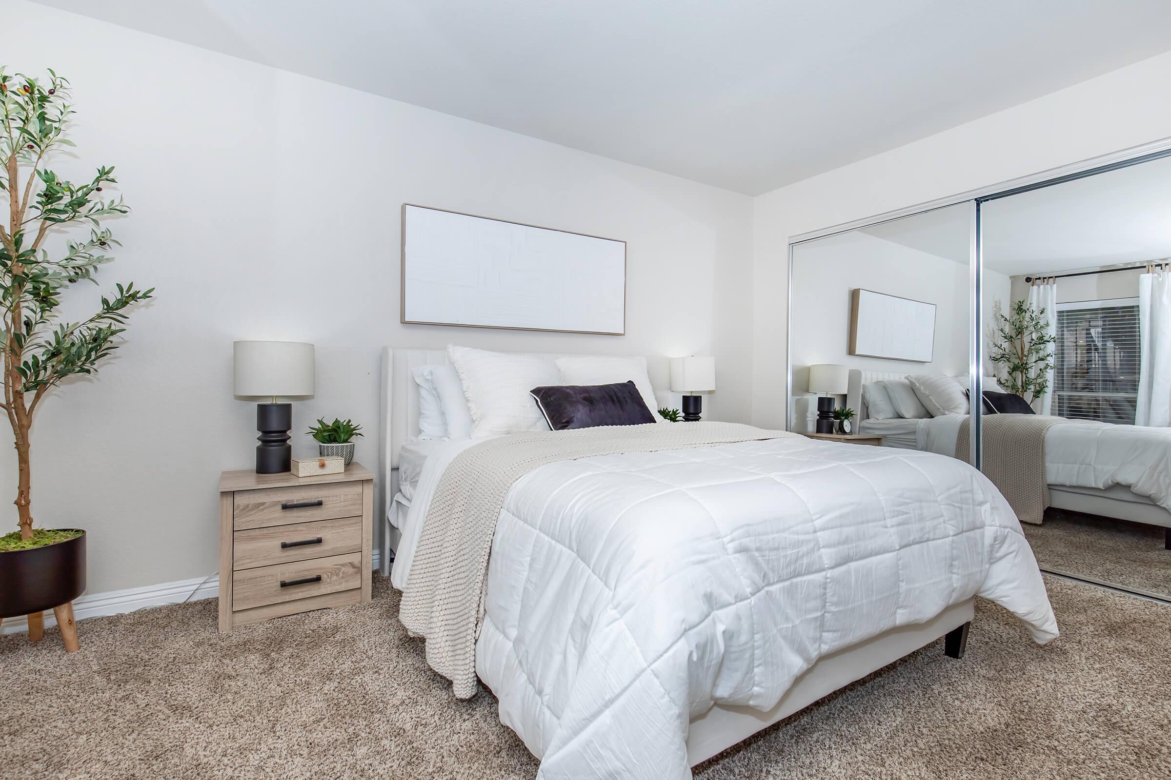 A cozy bedroom featuring a neatly made bed with white bedding, two bedside lamps, a nightstand, and a decorative plant. There is a large mirror reflecting the room and light-colored walls, creating a bright and inviting atmosphere. The carpet adds warmth to the space.