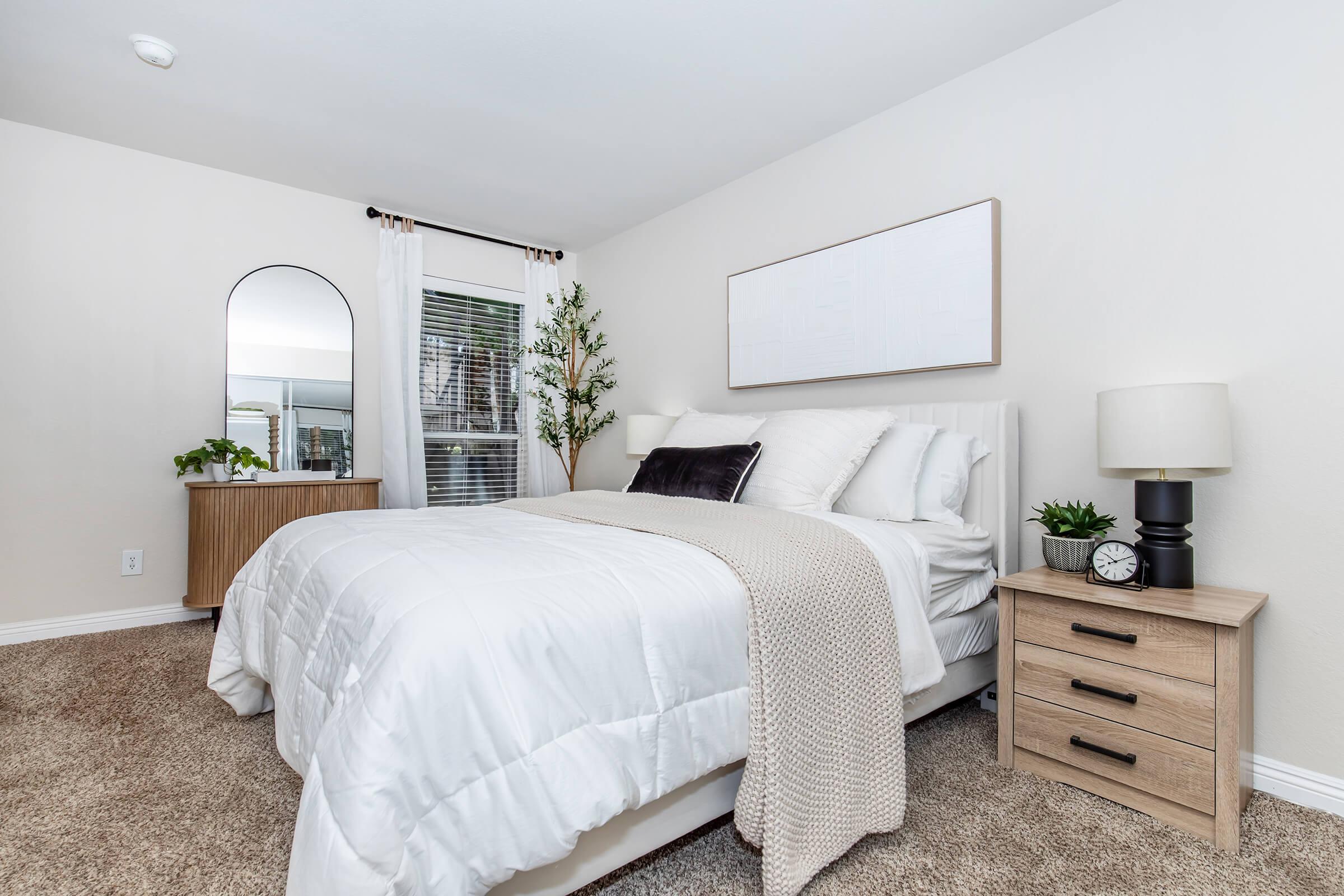 A cozy bedroom featuring a large bed with white bedding and decorative pillows. A mirror and nightstand are positioned beside the bed, along with a small plant. The walls are painted a soft color, and a window with blinds allows natural light to enter the space, complemented by a potted plant in the corner.