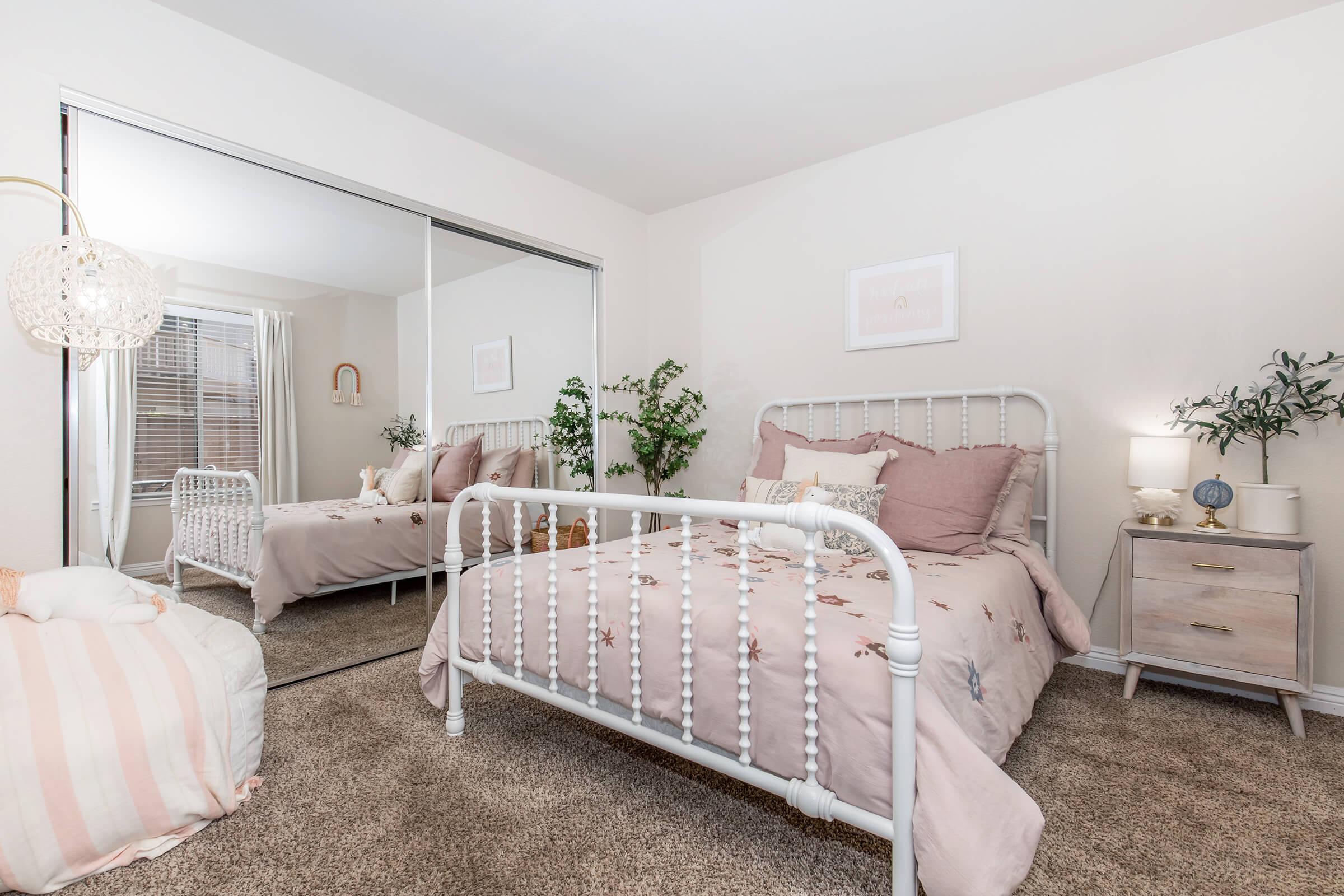 A cozy bedroom featuring a white metal bed with a pink embroidered duvet, decorative pillows, and a mirrored closet. Natural light floods the room, highlighting a potted plant, a bedside lamp, and a small nightstand. Soft beige carpet adds warmth to the space.