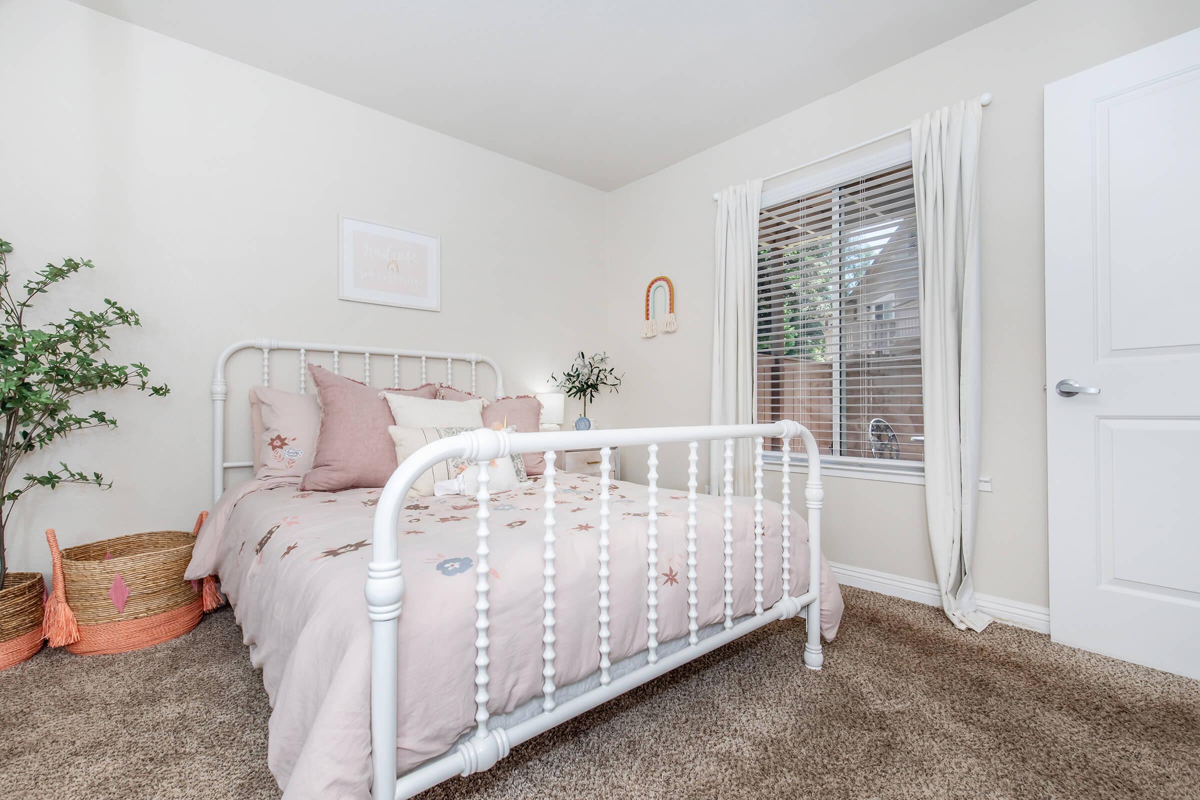 A cozy bedroom featuring a white metal bed with pink and neutral bedding, surrounded by a soft carpet. There's a window with light curtains allowing natural light in, and a small plant and decorative items add a touch of warmth. Two woven baskets are placed beside the bed, enhancing the room's inviting atmosphere.